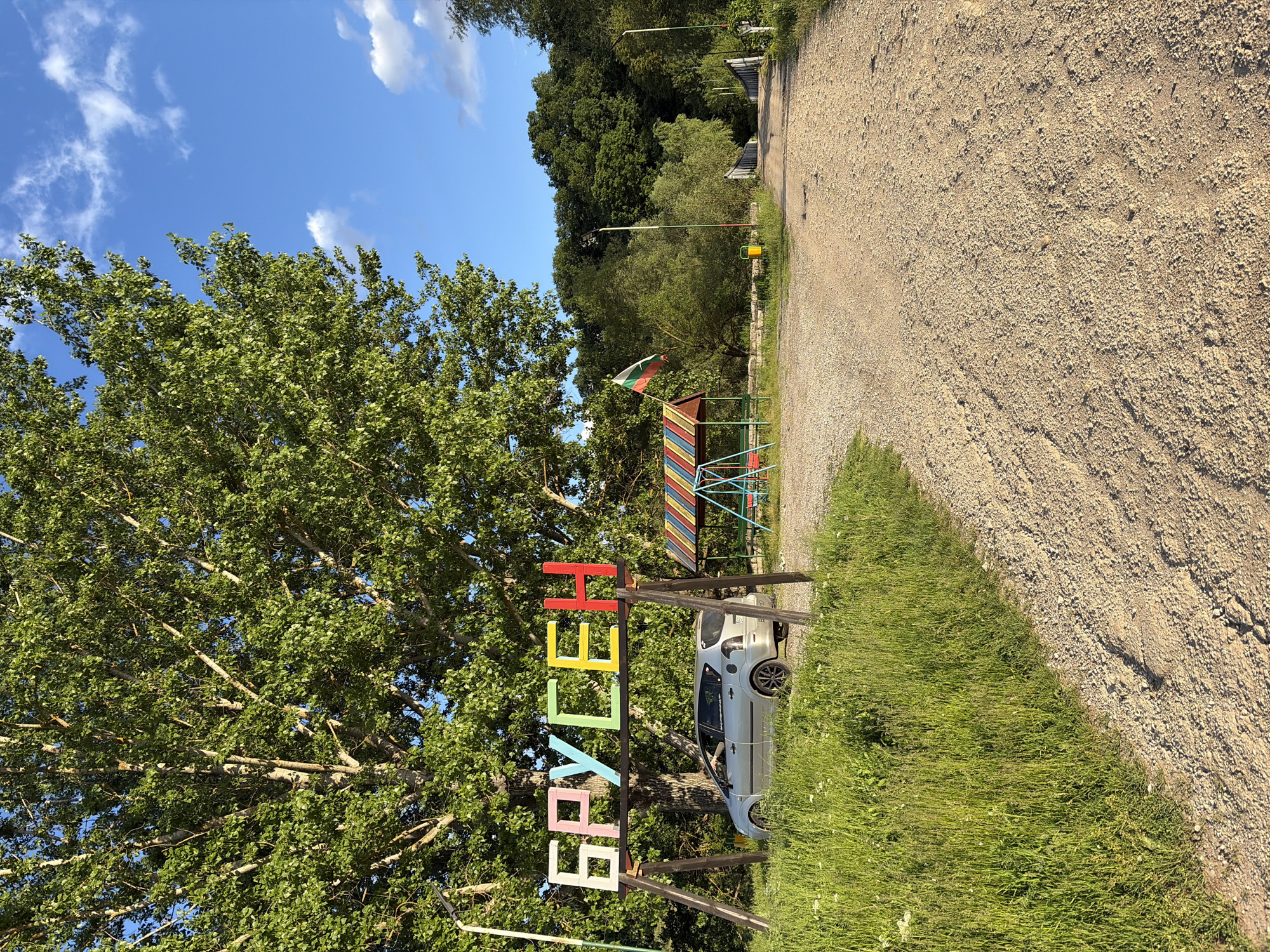 The image shows a dirt road leading into a forested area, presumably a park. On the left, there's a large tree and a sign that reads "БРУСЕН" (Brusen in Cyrillic alphabet) with each letter painted a different color. A parked car is near the sign. A colorful children's climbing structure is behind the sign. The flag of Bulgaria is atop the climbing structure. The sky is blue with some scattered clouds. Based on the sign "БРУСЕН" and context, this is in or near the village of Brusen, Bulgaria.