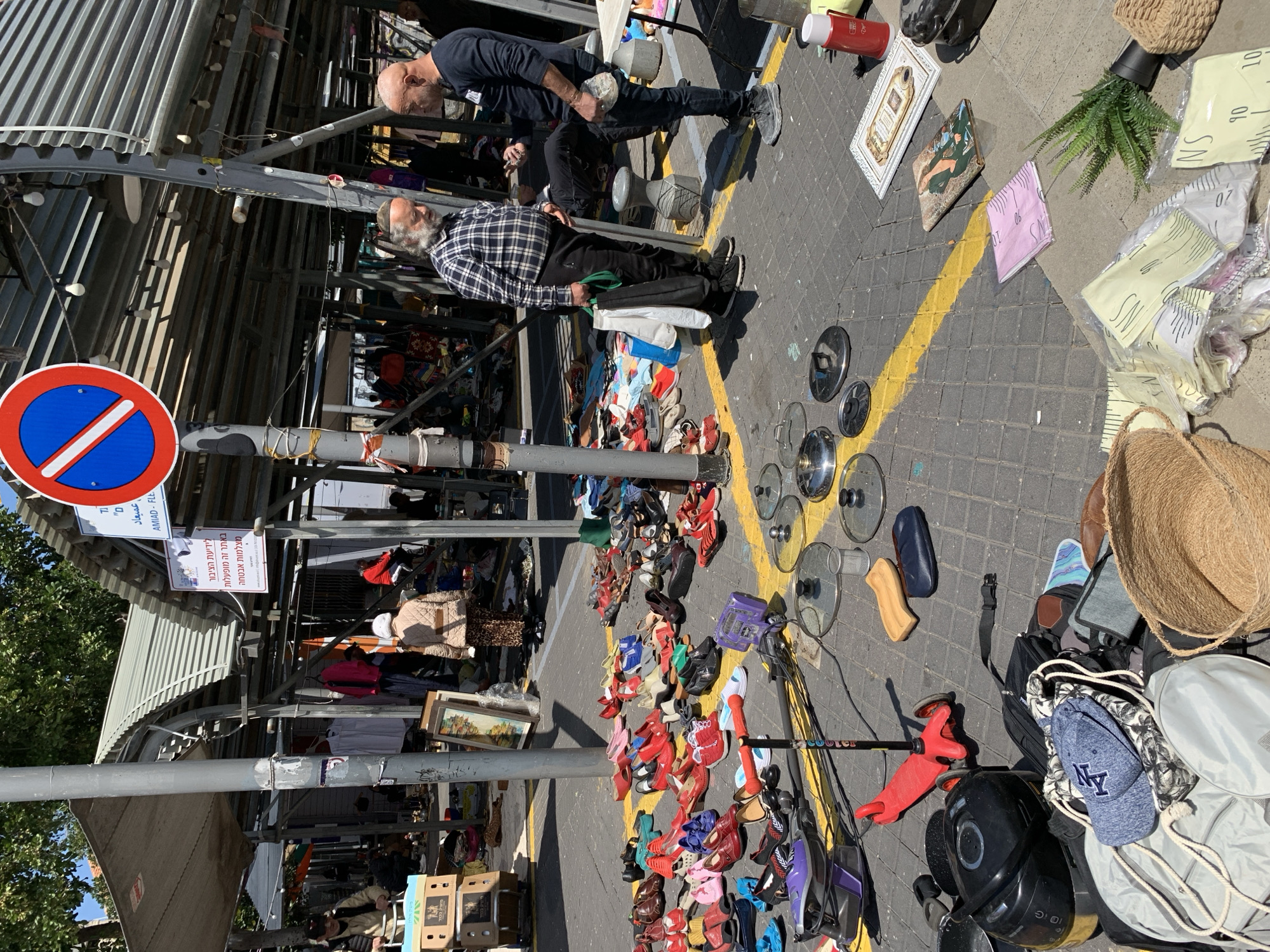 The scene depicts a street market in Tel-Aviv, Israel. The market is under a metal structure supporting lights. The pavement is marked with yellow lines and is covered with various items for sale, including shoes, cookware, books, plants, and bags. Two men are positioned near the edge of the market. One man is sitting on a stool while the other is standing nearby. The men appear to be shoppers. To the left, there is a blue and red "no parking" sign. Various signs in Hebrew are also attached to the metal structure.