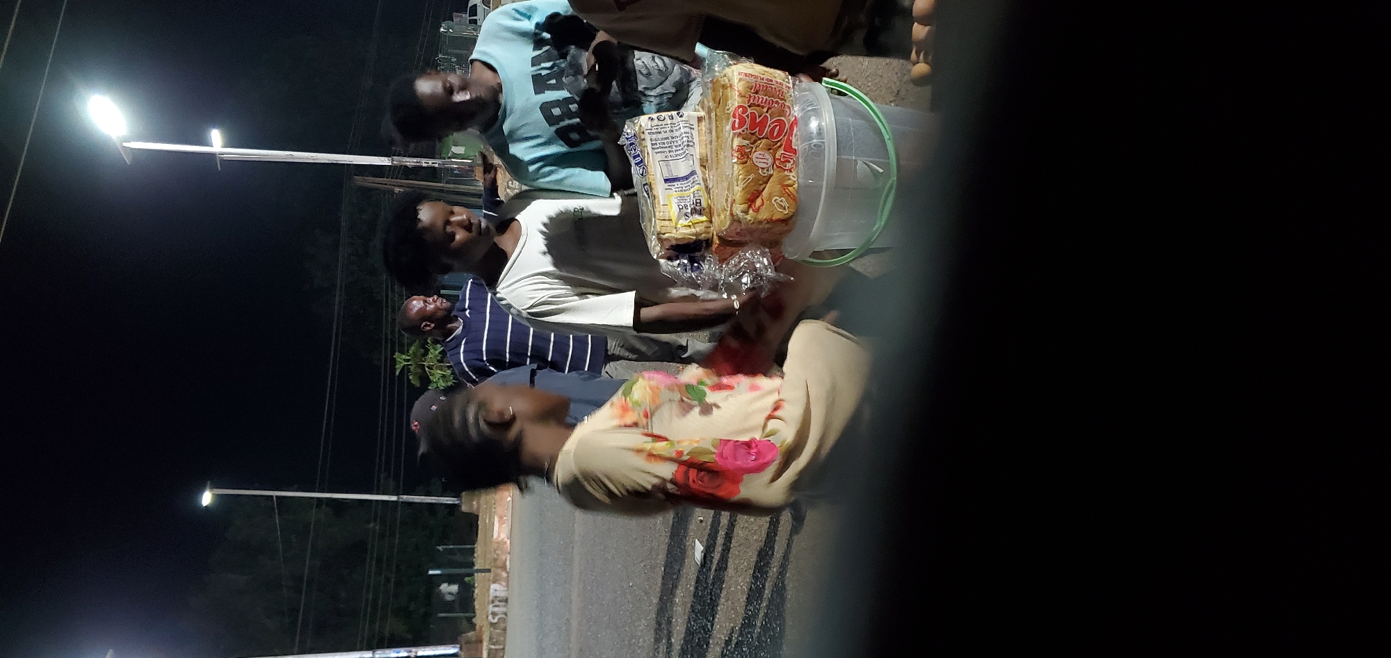 A nighttime scene depicts a group of individuals congregated around what appears to be a vendor's cart. The cart contains loaves of bread packaged in clear plastic, along with a clear plastic bucket with a green handle. The vendor, or person tending to the cart, is wearing a white shirt. A person in a light blue sweatshirt appears to be looking at the items on the cart. Another individual stands nearby wearing a white and blue striped shirt. In the foreground, a person wearing a light-colored shirt with a floral pattern is visible. The setting appears to be a city street, based on the presence of a street lamp and a paved road. The location is Unknown, Unknown.
