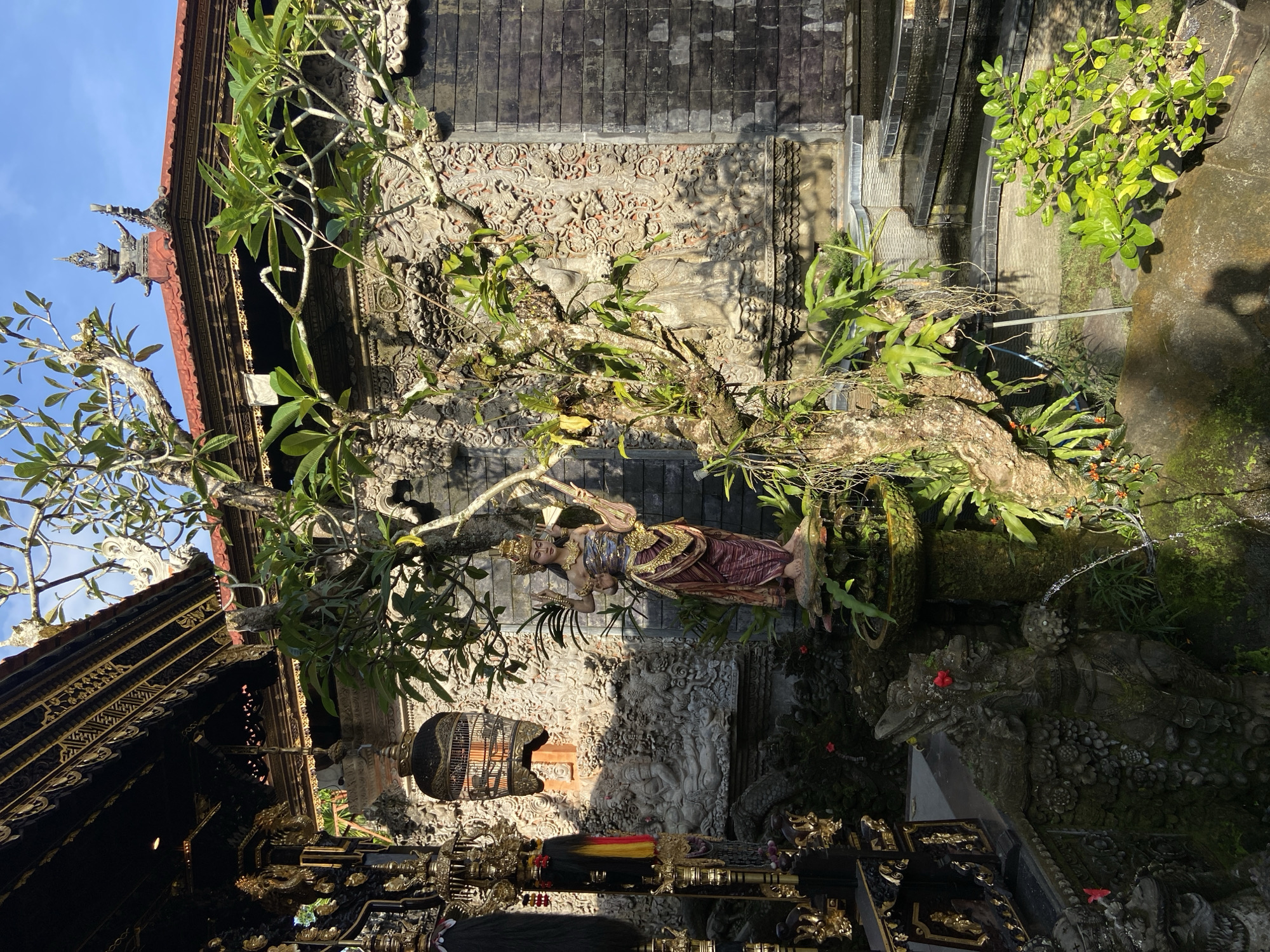 The image shows an outdoor scene in the Ubud District, Indonesia, featuring a Balinese temple complex. Dominating the center is a life-sized statue of a dancer adorned in traditional Balinese attire, integrated with a tree that has foliage. Intricate stone carvings are visible on the temple walls, featuring depictions of figures and ornate patterns. A black birdcage hangs near the left of the image next to a traditional roof that is ornate with golden patterns. Below is a display of objects that appears to be a market or offering area. The background consists of lush greenery and water feature, which adds to the atmosphere of the complex. The lighting indicates a sunny day, with shadows.