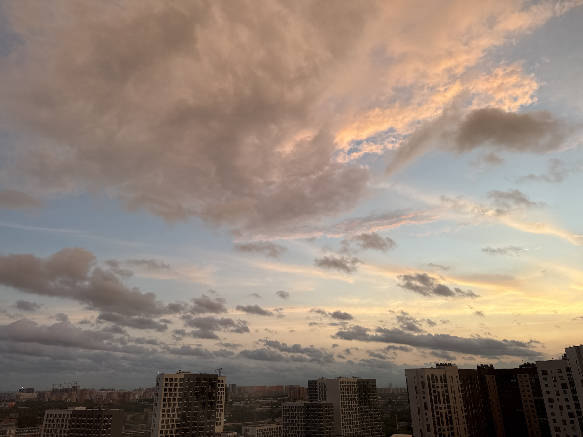 This is an aerial, wide-angle photograph taken in the city of Lyubertsy, Russia, during a sunset. The sky dominates the upper two-thirds of the frame, filled with an array of clouds in varying shades of pink, orange, and gray. The clouds are illuminated by the setting sun, creating a dramatic and colorful display. The lower third of the image shows a cityscape, characterized by numerous tall, multi-story apartment buildings. The buildings are constructed with a combination of light-colored facades and darker accents, featuring numerous windows. A few construction cranes are visible in the distance amongst the buildings, suggesting ongoing development. The overall impression is one of a modern urban environment under a vibrant, colorful sky.