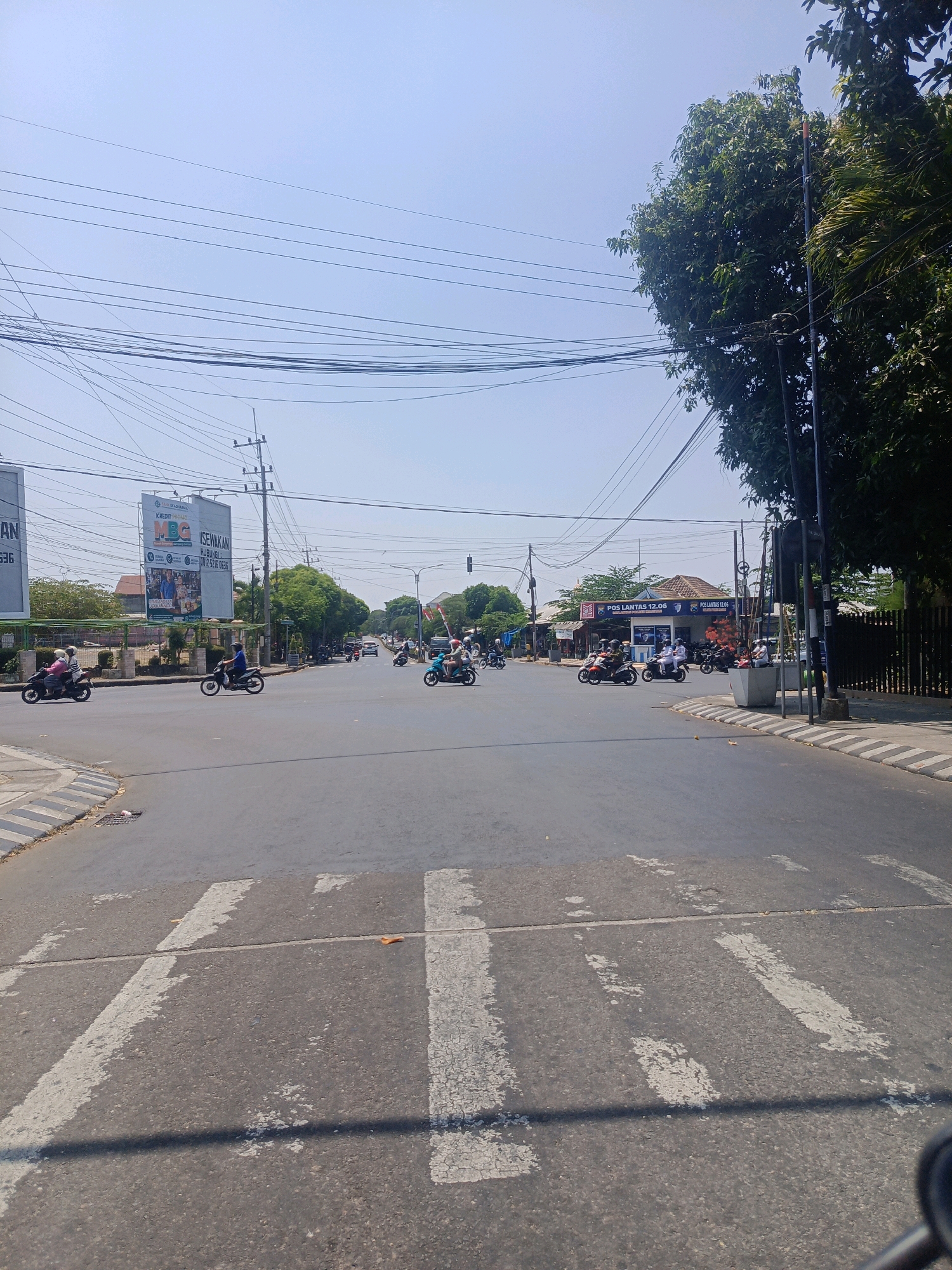 The image depicts a street intersection in Sukowinangun, Indonesia. A paved road with white pedestrian crossing lines runs through the center of the frame. Numerous motorcycles are present on the road, with riders in motion. Some motorcycles are turning, while others are proceeding straight. In the background, there are buildings, including one with a sign that reads "POS LANTAS 12.06". Several trees and utility poles with a complex network of overhead wires are visible, indicating an urban environment. The sky is clear and blue, suggesting a sunny day. The overall scene conveys a typical street view with moderate traffic activity.