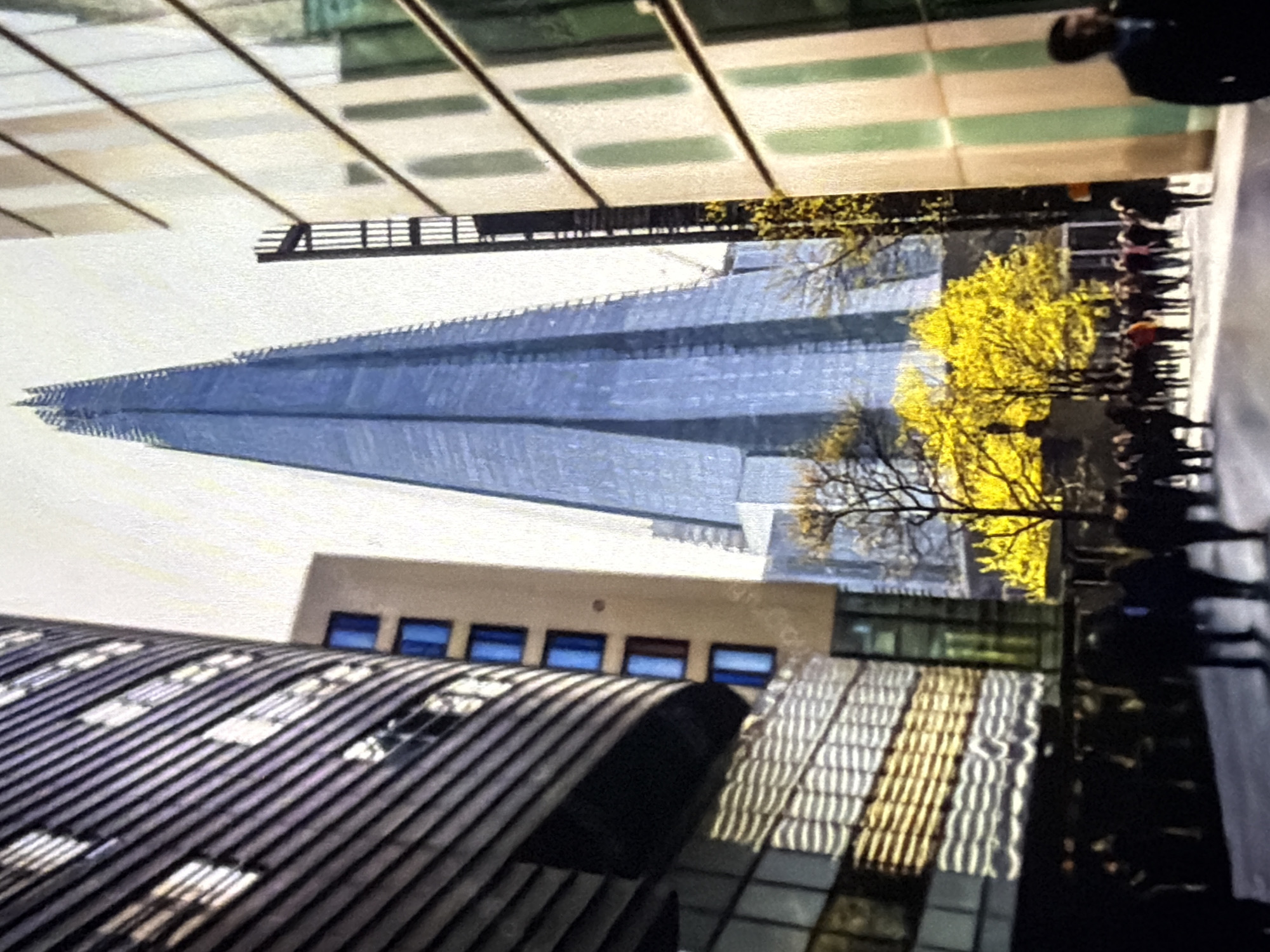 The image captures an urban scene in **London, United Kingdom**, prominently featuring The Shard skyscraper.
In the background, the tall, slender, glass-clad structure of The Shard dominates the center, rising towards a bright, featureless sky, reflecting a bluish hue. To the left, a large, multi-story building with a dark, horizontally banded or paneled facade curves into the foreground. Behind it, a lighter, rectangular building with a series of vertically stacked square windows featuring bright blue frames is visible. On the right, another multi-story building with a pale facade displaying vertical greenish-white sections extends into the foreground. Between this building and The Shard, a dark, ladder-like structural element is present.
In the mid-ground, beneath The Shard and the surrounding buildings, several deciduous trees with distinct clusters of bright yellow foliage are visible, suggesting an autumnal setting. The lower half of the image depicts a paved pedestrian area, where a crowd of approximately 30-40 individuals, mostly indistinct due to distance and soft focus, are walking in various directions, primarily moving towards the background. Most pedestrians appear to be dressed in dark clothing. A man in a dark suit or coat is partially visible in the lower-right foreground, facing away from the viewer. The overall lighting is bright and diffuse, indicative of an overcast day.