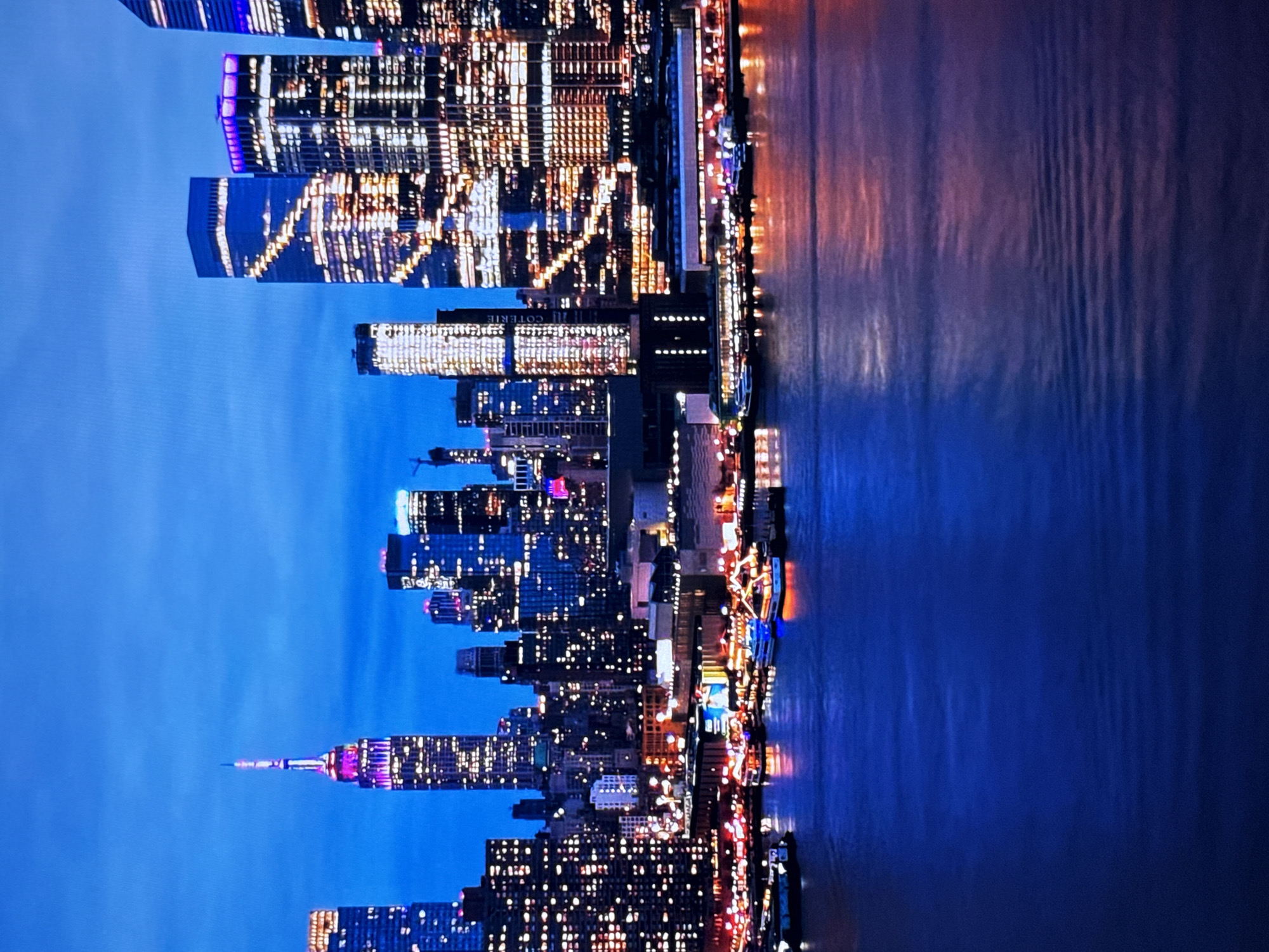 The image shows a panoramic view of a city skyline at dusk, illuminated by the lights of numerous buildings and a body of water in the foreground. The skyline features a variety of skyscrapers with many windows reflecting the ambient light, creating a dazzling effect. One prominent skyscraper is lit in purple and white, suggesting it's a landmark. Several boats are visible on the water, some adorned with colorful lights, hinting at a nighttime event or a busy harbor. The sky is a deep blue, transitioning from twilight to night. The water reflects the city lights, creating streaks of color on its surface. This scene depicts a vibrant urban landscape at night, likely a major city with a waterfront. The location is not Burgas, Bulgaria; it is New York City.