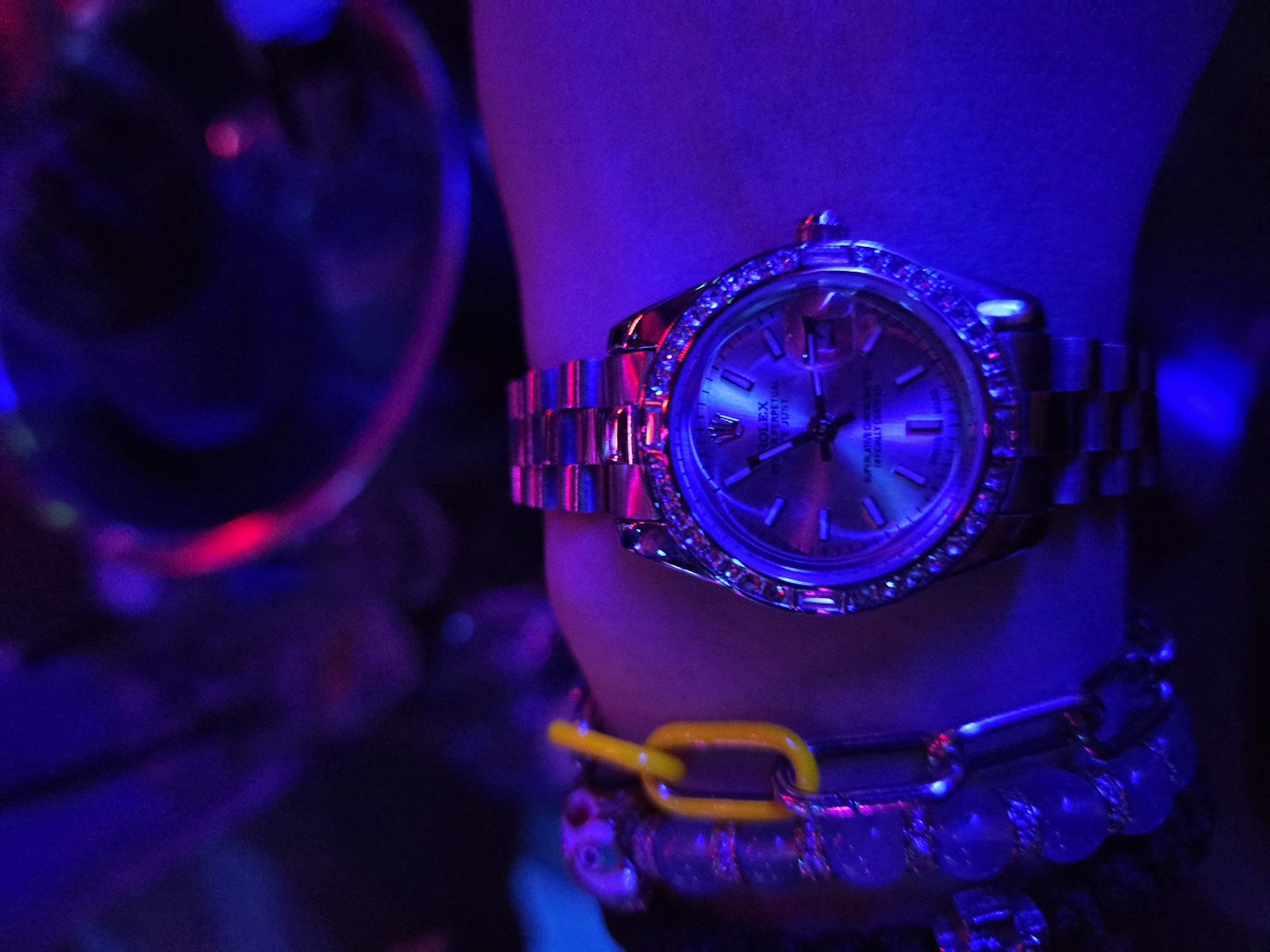 A wrist adorned with a Rolex watch and several bracelets is presented in a setting bathed in purple and blue lighting. The Rolex watch, featuring a silver band and a face encircled by gemstones, is the focal point. Beneath it, a collection of bracelets is visible, including one with a yellow chain link. The overall atmosphere suggests a nighttime or low-light environment. The location cannot be determined from the image.