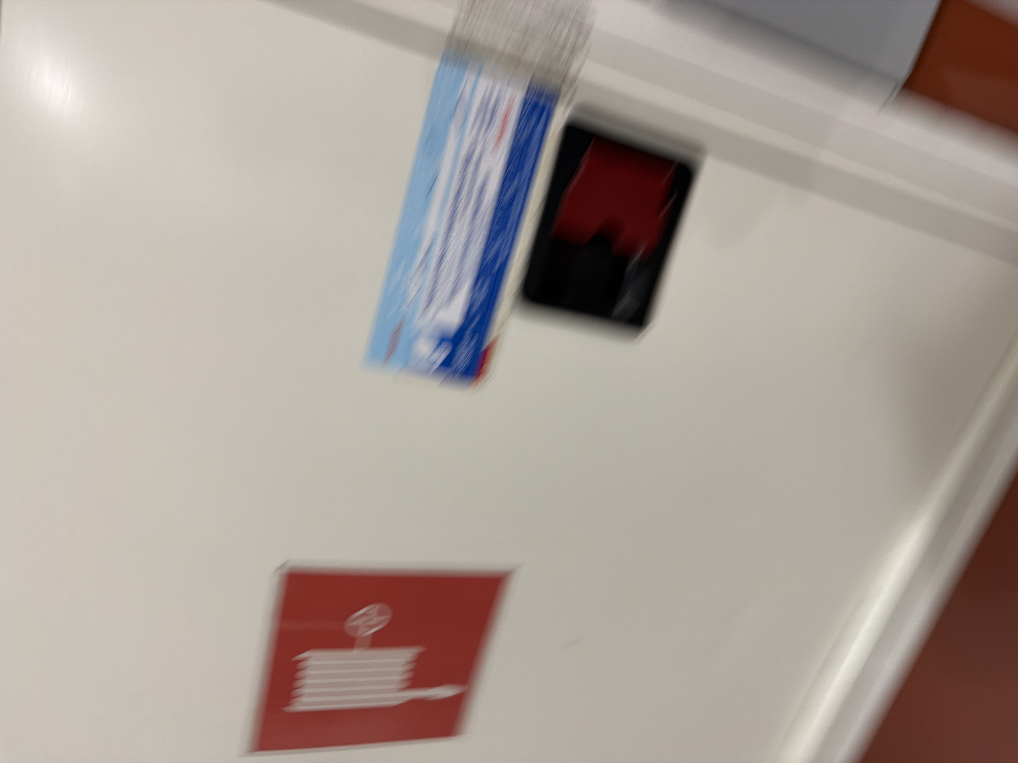 The image shows a white wall with two objects affixed to it and a portion of a red floor at the bottom. On the left, a red square sign with a white icon of a coiled hose and a nozzle, likely indicating a fire hose reel, is visible. To the right of the fire hose sign, a long, rectangular object with blue and white graphics, possibly packaging or a sign, is mounted horizontally. Below this, a black rectangular object with a red shape visible within it is attached. The overall image is blurry and appears to be taken from a handheld camera, suggesting movement during the photograph. The lighting is diffuse, and the environment appears to be indoors, possibly a public building or corridor, given the signage. The context of Sofia, Bulgaria, is not visually ascertainable from the provided image.