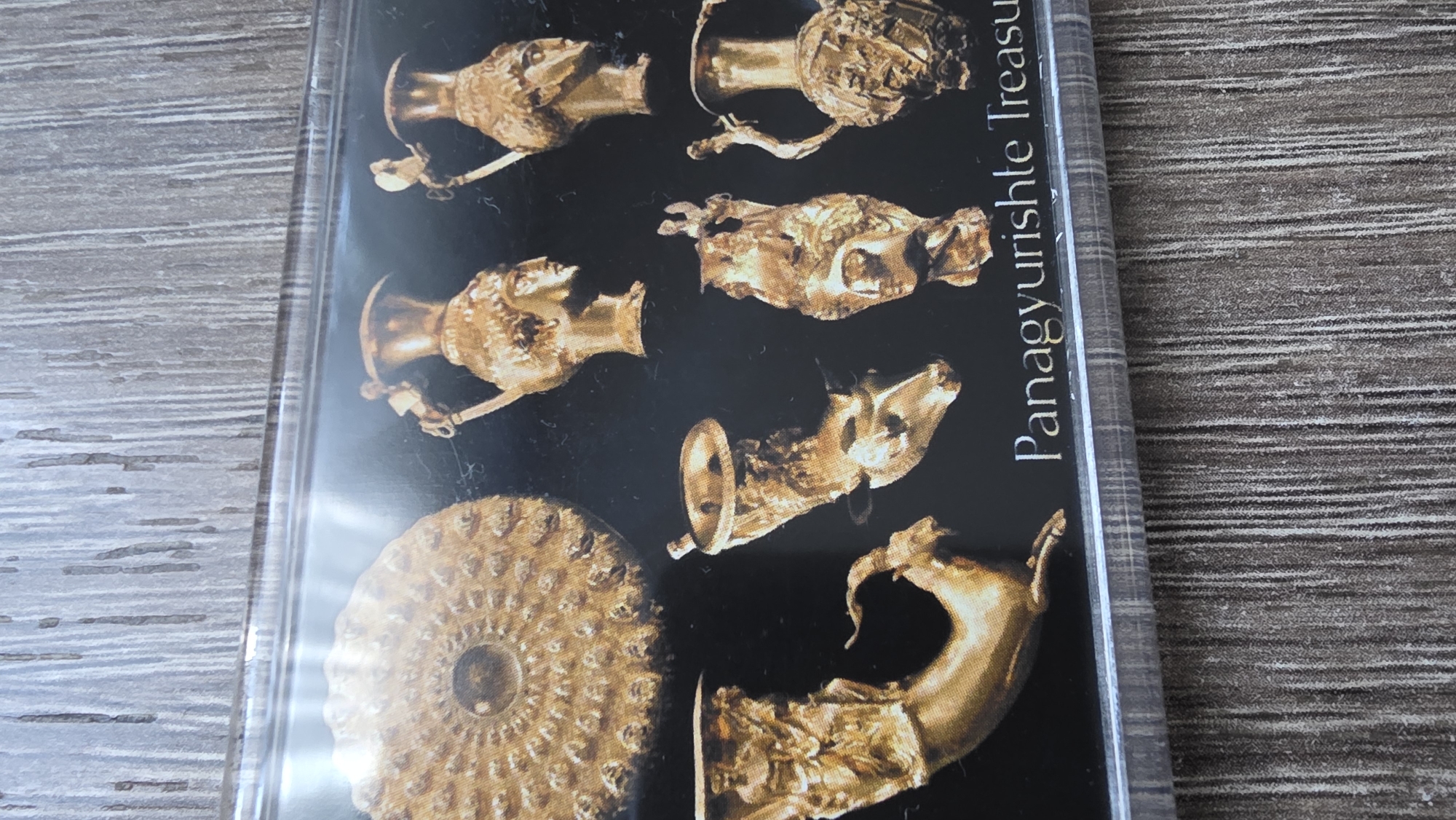 The image shows the cover of a book titled "Panagyurishte Treasure." The cover depicts several golden artifacts against a black background. These include rhytons (drinking vessels) and other gold objects. The book is resting on a wooden surface. There is no mention of Blagoevgrad, Bulgaria, in the immediate image content.