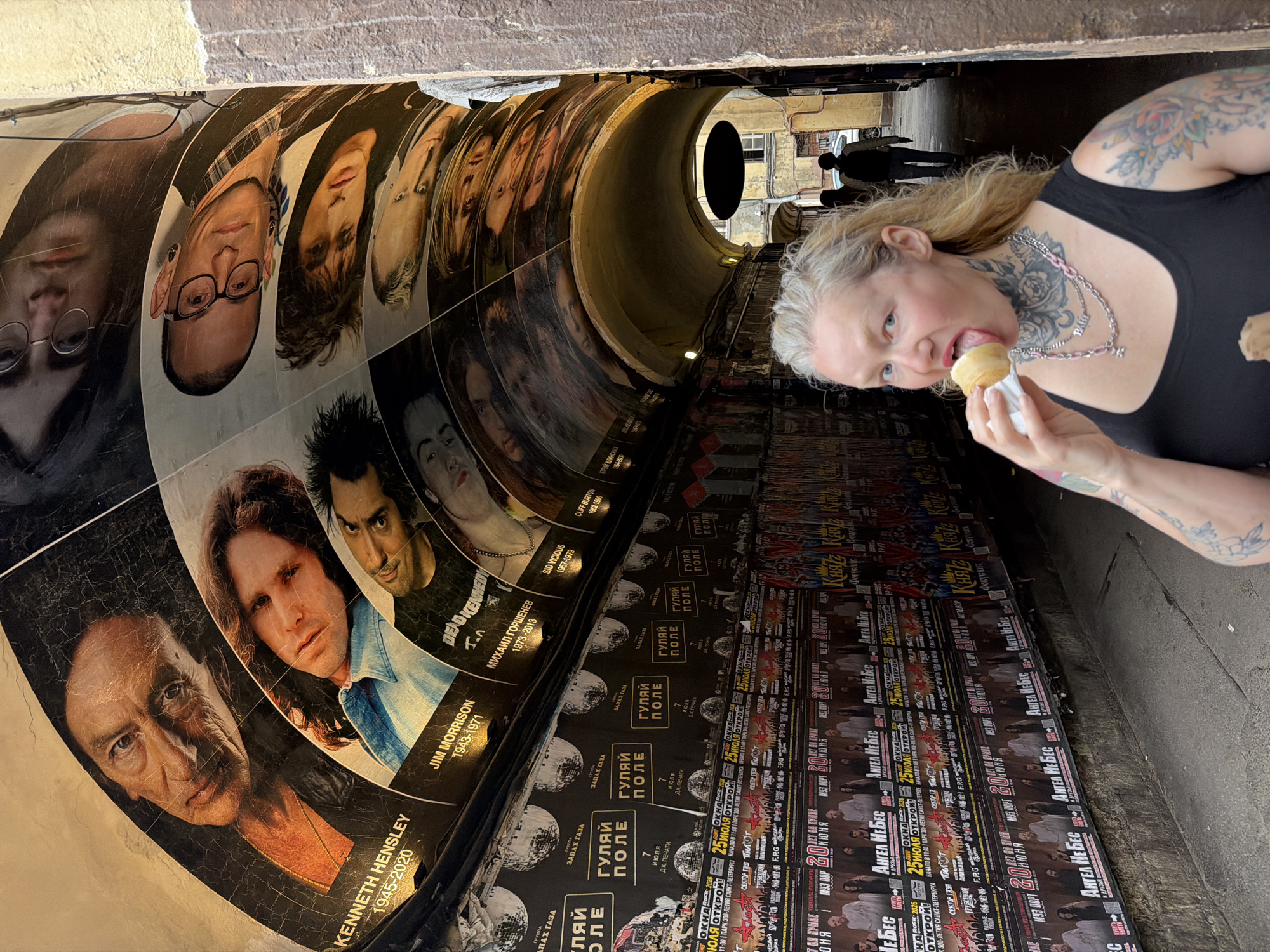 The image captures a street scene in Saint Petersburg, Russia, specifically within an arched passageway adorned with posters and murals of musicians. A blonde woman with extensive tattoos on her neck, arms, and chest is in the foreground, holding an ice cream cone to her mouth and licking it. She is wearing a black tank top and multiple necklaces, one of which has the word "BABY" on it.
The arched ceiling of the passageway is decorated with large portraits of musicians, including Kenneth Hensley (1945-2020) and Jim Morrison (1943-1971), along with other faces. The walls on either side of the passageway are covered in layers of posters, some of which appear to be concert advertisements with Cyrillic text. These posters are densely packed and create a visually busy backdrop.
In the background, two individuals are walking away from the camera down the passageway. The environment appears to be an urban outdoor setting, with worn stone or brick textures on the sides of the passage. The lighting suggests it might be daytime, though parts of the passageway are in shadow. The overall atmosphere is urban and somewhat gritty, with the vibrant artwork providing a counterpoint to the weathered surfaces.