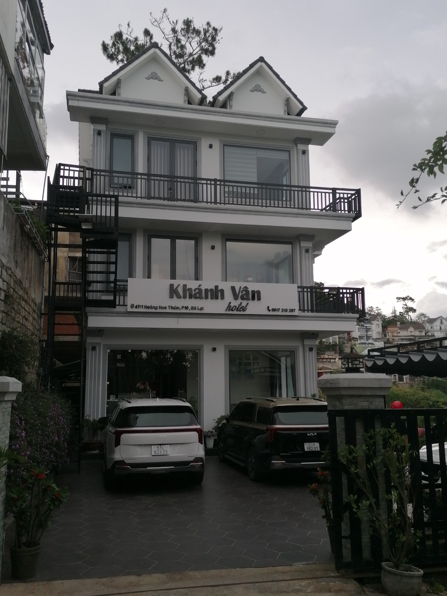 A three-story white building with black railings on the balconies and a black spiral staircase on the left side of the building is shown in Da Lat, Vietnam. A sign with "Khánh Vận hotel" is visible on the front of the building. Two vehicles, a white Kia and a black Kia, are parked in front of the building. The sky is overcast. The building appears to be a hotel or a guesthouse. The street number "47/1 Hoàng Hoa Thám, P10, Đà Lạt" is visible on the sign.