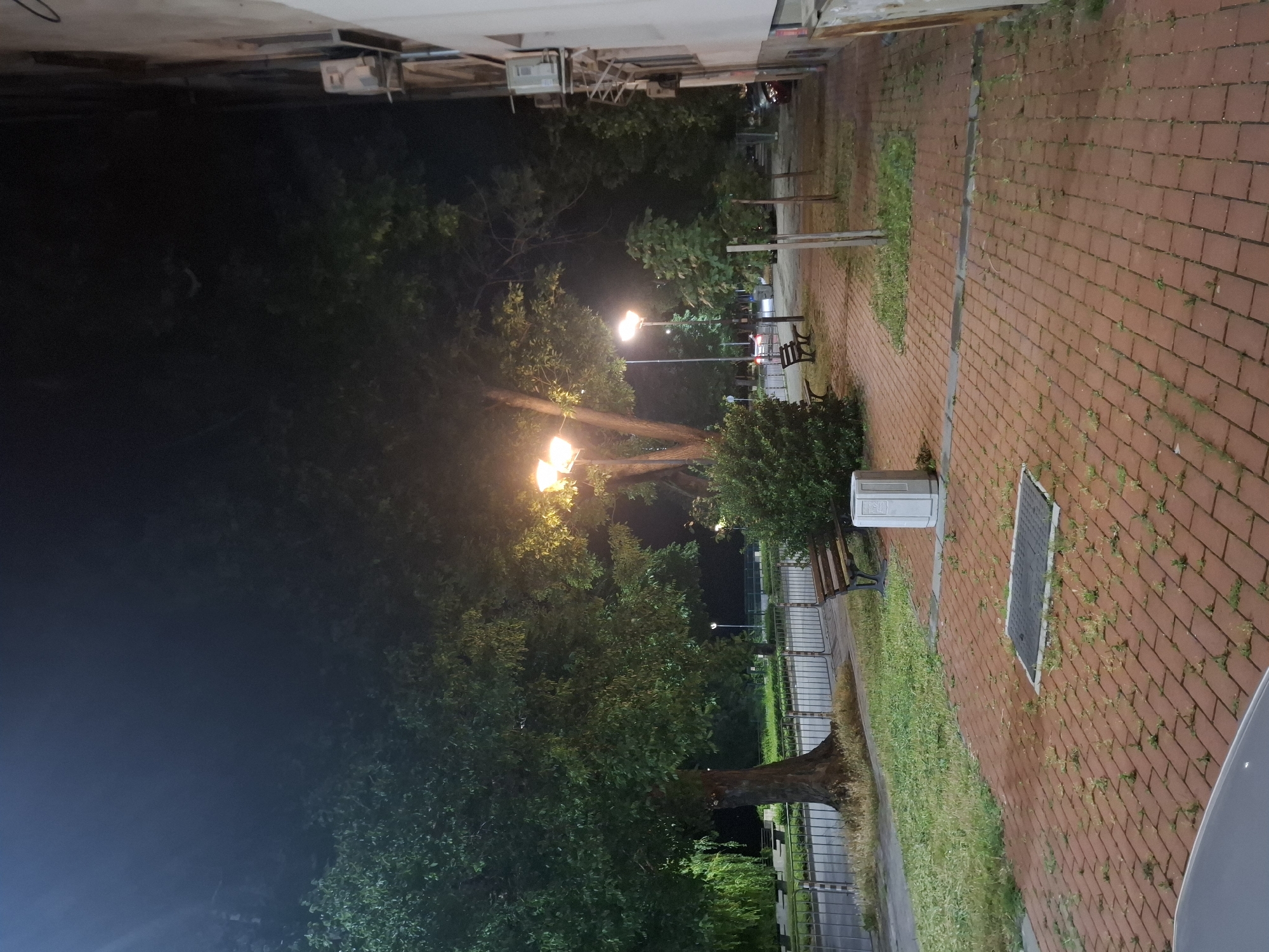The image depicts a nighttime scene on a sidewalk paved with red bricks. A large tree with lush green foliage stands to the left, its trunk thick and textured. To its right, a street lamp illuminates the area with a warm, yellow glow. A wooden bench is visible next to the tree, and further down the sidewalk, another bench is placed near a neatly trimmed bush. A cylindrical trash bin made of white concrete sits on the sidewalk. Beyond the sidewalk, a metal fence separates it from a grassy area and a road. Several smaller trees are planted along the sidewalk, and a building with an air conditioning unit mounted on its exterior wall is present on the right side of the image. The sky is dark, indicating it is nighttime.