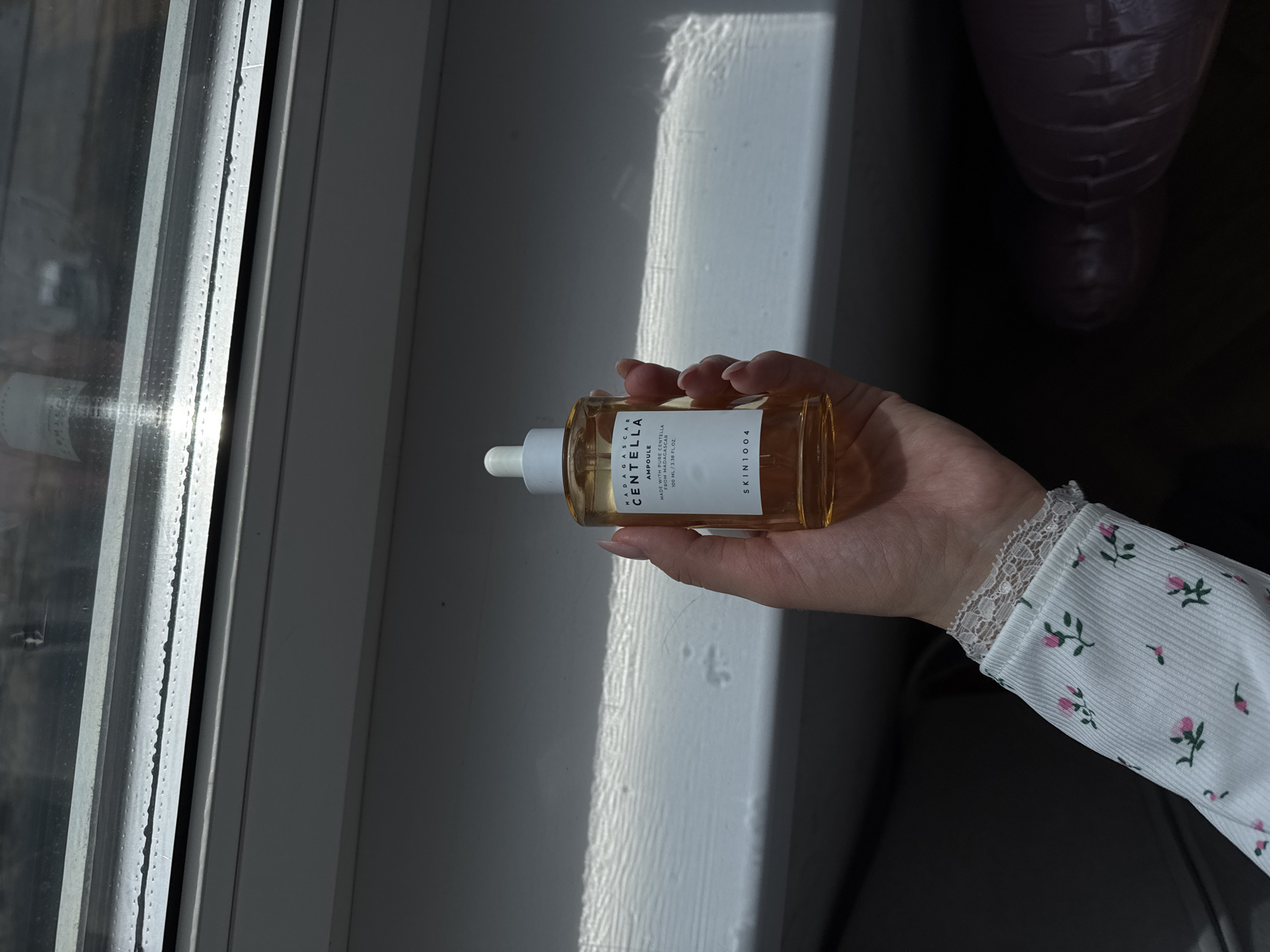 The image shows a bottle of SKIN1004 Madagascar Centella Ampoule held in a person's hand. The bottle is amber-colored with a white label and dropper. The hand is pale with manicured nails. The person is wearing a white shirt with a floral pattern and lace trim at the wrist. The background consists of a window frame and some blurred objects. The lighting suggests the photo was taken during the day.