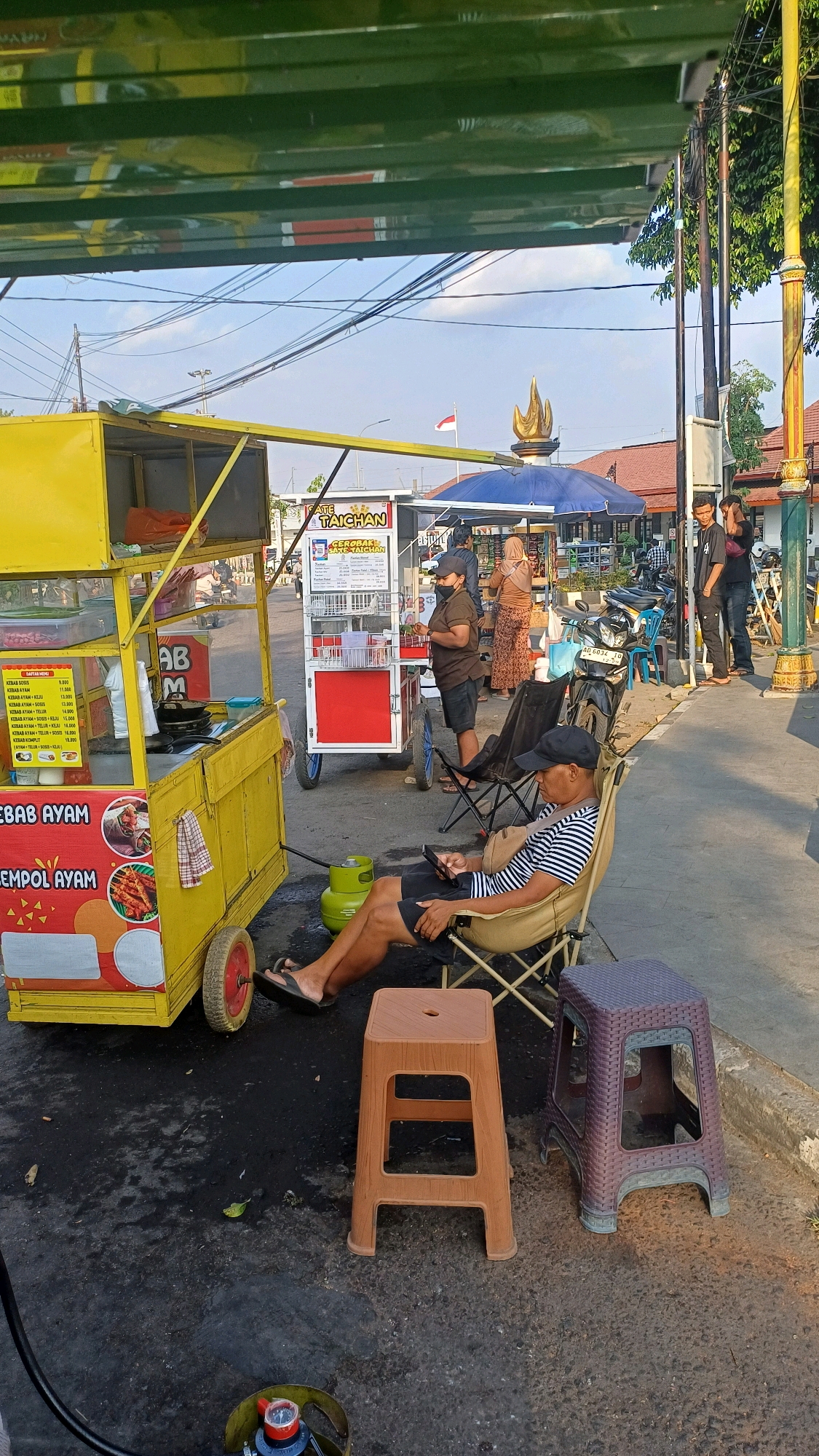 The image captures an outdoor scene, likely on a street, in Klaten, Indonesia. In the foreground, a man wearing a striped shirt and a black cap is seated in a beige camping chair, looking at his phone. He is wearing shorts and flip-flops. Beside him are two plastic stools, one brown and one purple. A yellow food cart, adorned with images of food items like chicken skewers, is to the left. A green gas cylinder is visible near the cart.
Behind the man, another food cart, this one white and red, displays a menu. A woman in a dark shirt and shorts, wearing a mask, stands by this cart, attending to customers. Further back, several people, including women in hijabs and men, are seen interacting. A blue umbrella is set up, providing shade. Motorcycles are parked in the background. The environment appears to be a street with a sidewalk. The sky is visible with some electrical wires overhead. The overall atmosphere suggests a street food vendor area.