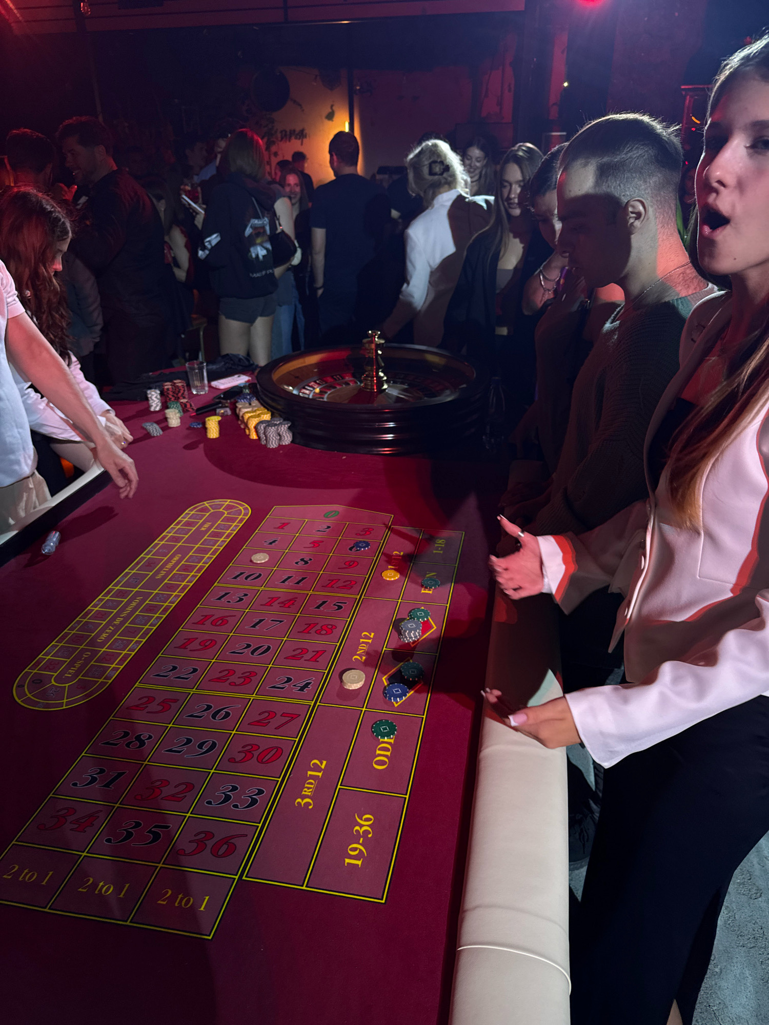A group of young adults are gathered around a roulette table in a dimly lit indoor space, possibly a club or event venue, in Rostov-on-Don, Russia. The roulette wheel is visible on the right side of the image, and the betting layout of the table is in the foreground. Several people are standing and looking at the table, with some appearing to place bets or watch the game. Chips and a glass are on the table. The atmosphere appears to be festive and social.