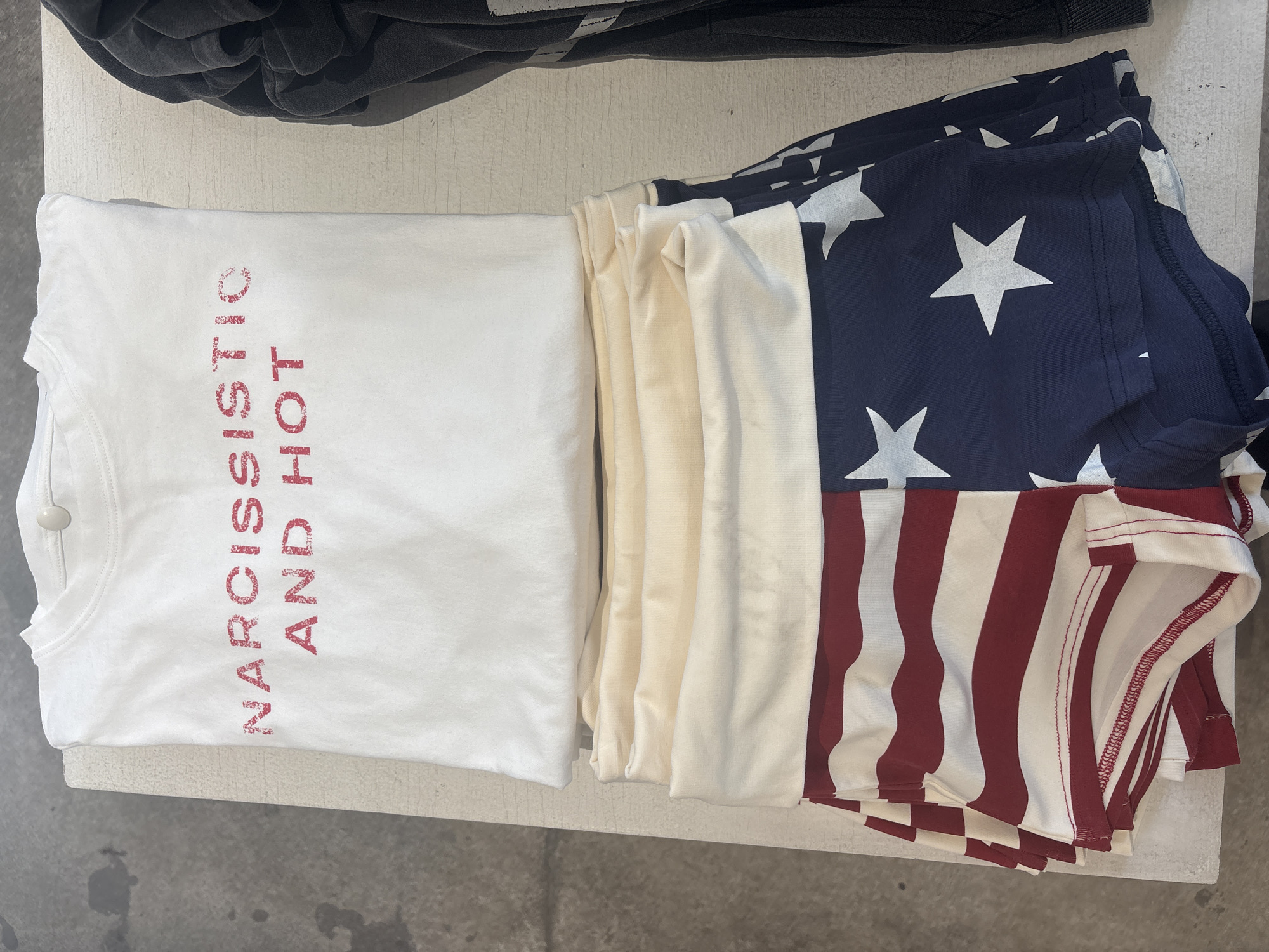 The image shows a stack of folded t-shirts on a light-colored surface. From left to right, the first shirt is white with red lettering that reads "NARCISSISTIC AND HOT" vertically. The next few shirts appear to be off-white or cream colored. The shirt on the far right is decorated with the American flag. There is also dark clothing in the upper portion of the image. The location is not discernible from the given image and therefore no assumptions can be made about the location being Moscow, Russia.