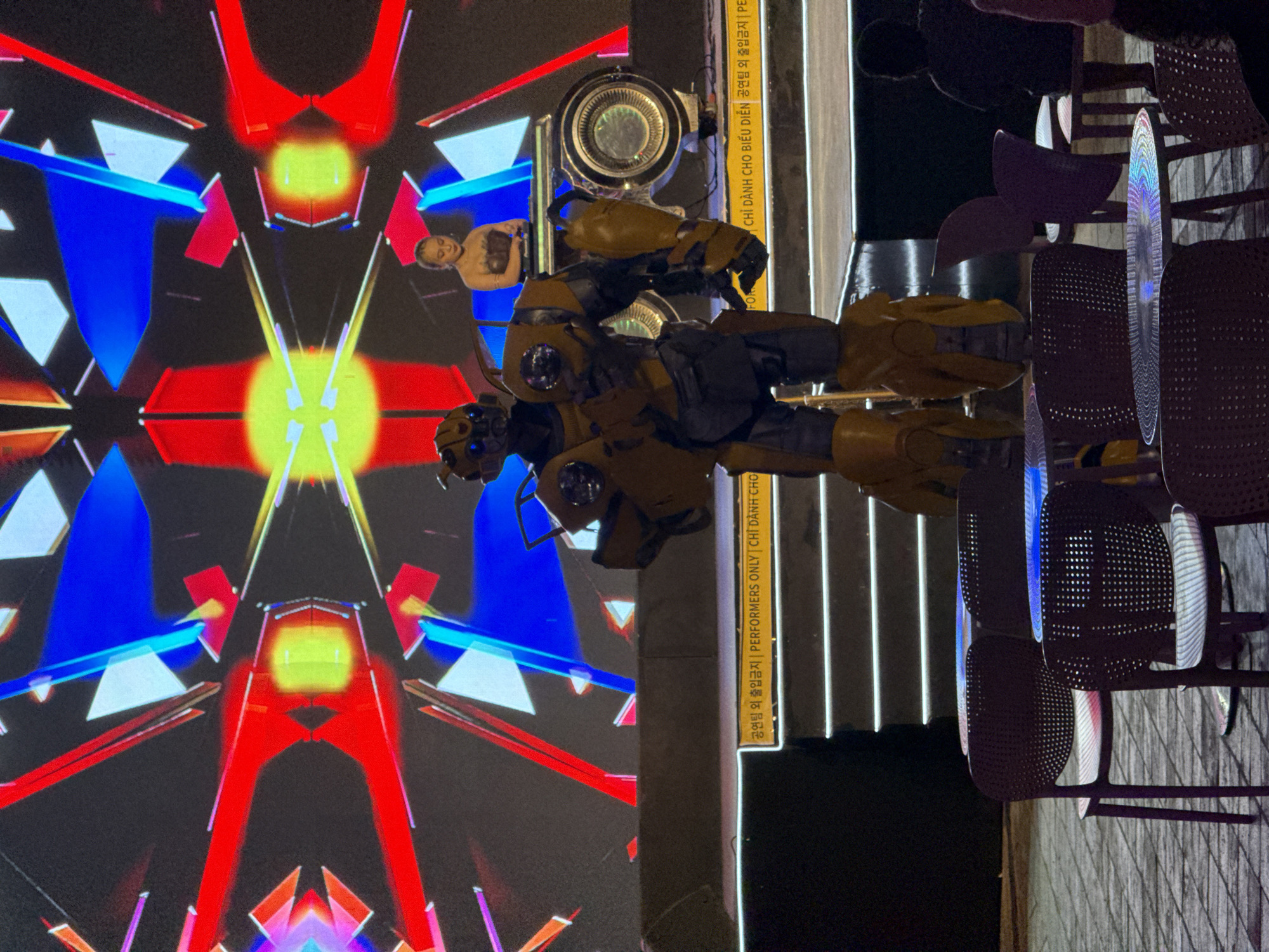 An evening outdoor scene in Đà Nẵng, Vietnam, features a large, detailed figure resembling Bumblebee from the Transformers franchise, standing prominently. The robot, primarily yellow and dark grey with glowing blue eyes, is positioned in front of a dazzling, symmetrical abstract light display. This background projection showcases vibrant geometric shapes in red, blue, yellow, and white, creating a dynamic visual effect. Partially visible within the background display, to the left of the robot's head, is a human figure, possibly a performer, suggesting an interactive or projected element of the show.
To the right of the robot, several dark, perforated chairs are stacked or arranged, implying a seating area for an audience. Above the robot, a circular light fixture is visible. A vertical column or beam separates the illuminated backdrop from the chairs.
Visible text on a vertical sign, appearing in both Korean, English, and Vietnamese, reads: "공연팀 외 출입금지 | PERFORMERS ONLY | CHỈ DÀNH CHO BIỂU DIỄN," which translates to "No entry except for performance team | Performers Only | For Performers Only." This indicates a performance area restricted to staff, suggesting an event or show is taking place. The overall ambiance is lively and artistic, with a focus on light and performance.