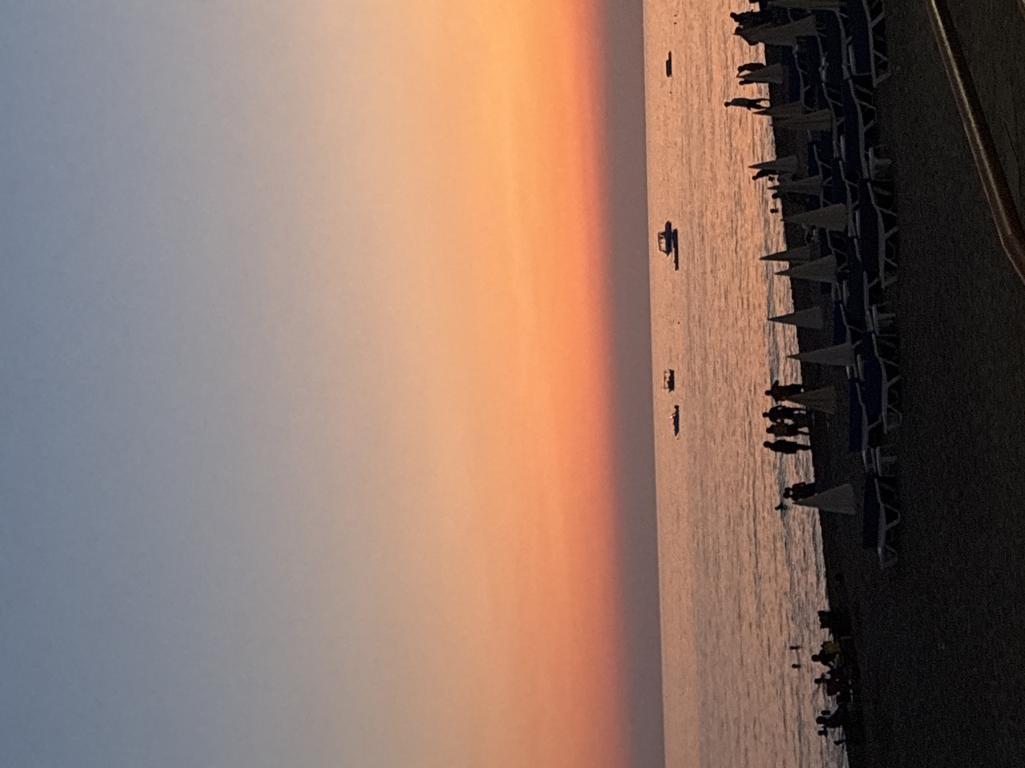 The image depicts a sunset scene at a beach in Alanya, Turkey. The sky transitions from a pale blue at the top to a vibrant orange and then a soft pink closer to the horizon. The sea is calm, with a few boats visible in the distance. On the shore, there are several beach umbrellas and lounge chairs. A group of people are seen walking along the water's edge and some are wading in the sea. The overall atmosphere suggests a relaxed evening at a popular tourist destination.