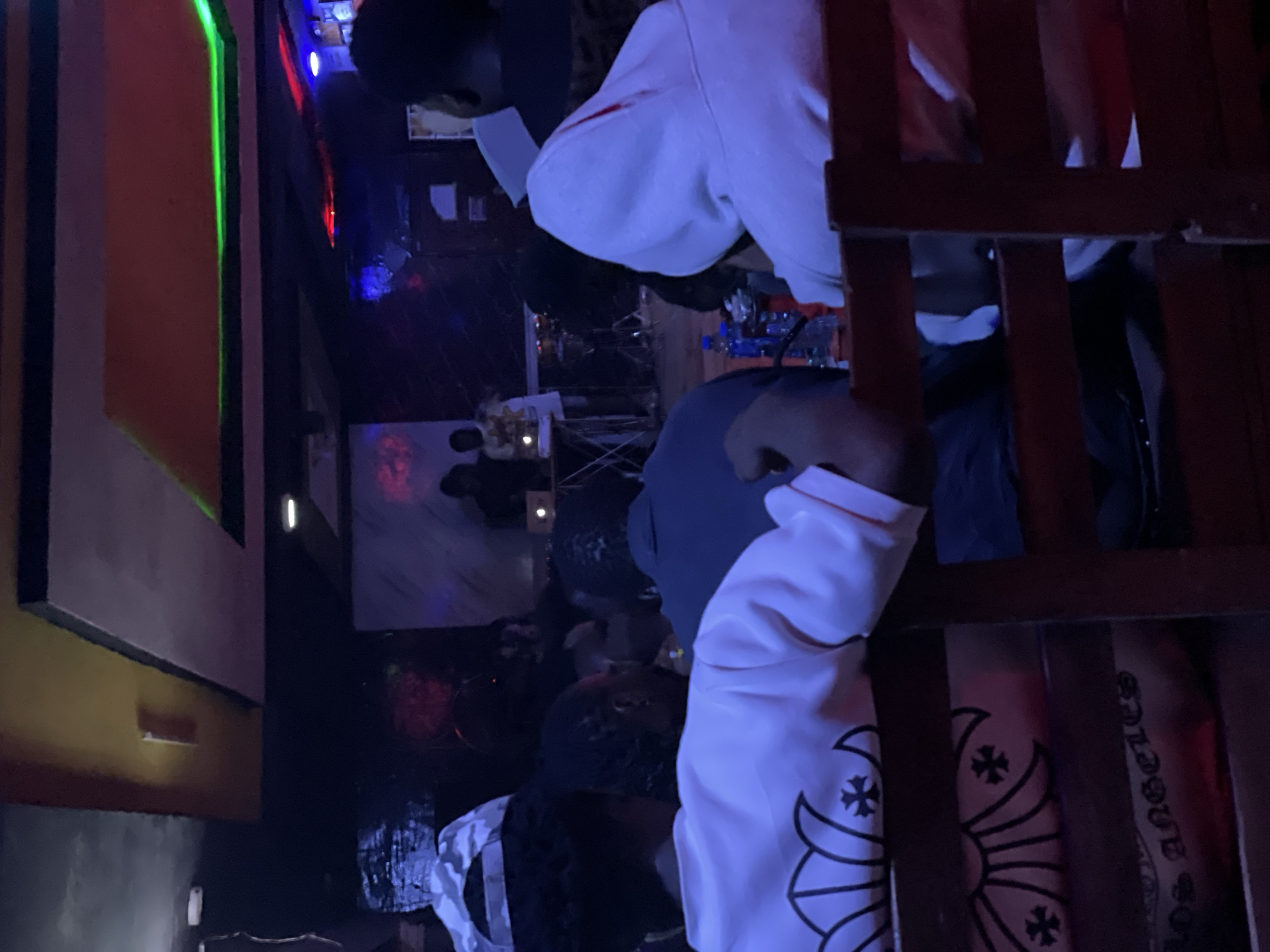 The image shows a dimly lit indoor scene, possibly a club or lounge, with several individuals present. In the foreground, two individuals are seated, with their backs to the camera. The person on the left wears a white shirt with a distinctive black floral and cross design, and text that appears to say "Los Angeles." The person on the right wears a light-colored hooded sweatshirt.
In the middle ground, a group of people are gathered, some appearing to be interacting with a DJ setup or stage in the background. The environment is characterized by dark paneling on the ceiling with colorful LED lighting, creating a vibrant and possibly energetic atmosphere. The overall impression is of a social gathering taking place at night. The mention of Durimi, Nigeria is a contextual detail provided outside of the visual information within the image itself, and therefore cannot be directly verified or described from the image alone.