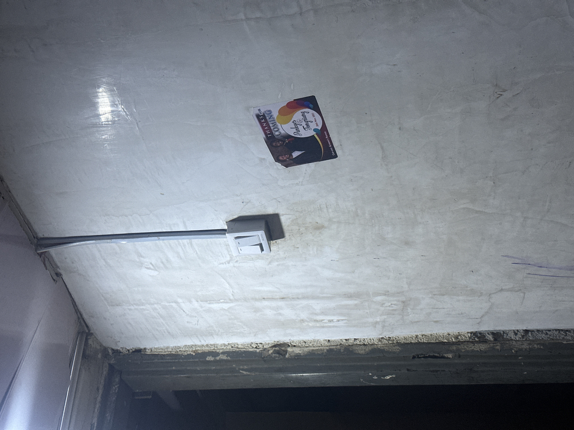 The image depicts an interior scene of a wall with a light switch and an invitation card attached. The wall is textured and appears to be unfinished or aged, with some visible cracks and marks. A metallic conduit runs vertically along the wall, terminating in a white electrical box housing a light switch. The invitation card, placed on the right side of the wall, features a photograph of a couple, balloons, and text that reads "THANKS FOR COMING Parkhyes & Tongliang 28th Nov. 2021". The bottom of the card also has text that reads "Cate.cre: Groom's Parent". The left side of the image shows a dark doorway or opening, with a rough, textured frame. The overall lighting suggests an indoor environment with artificial light.