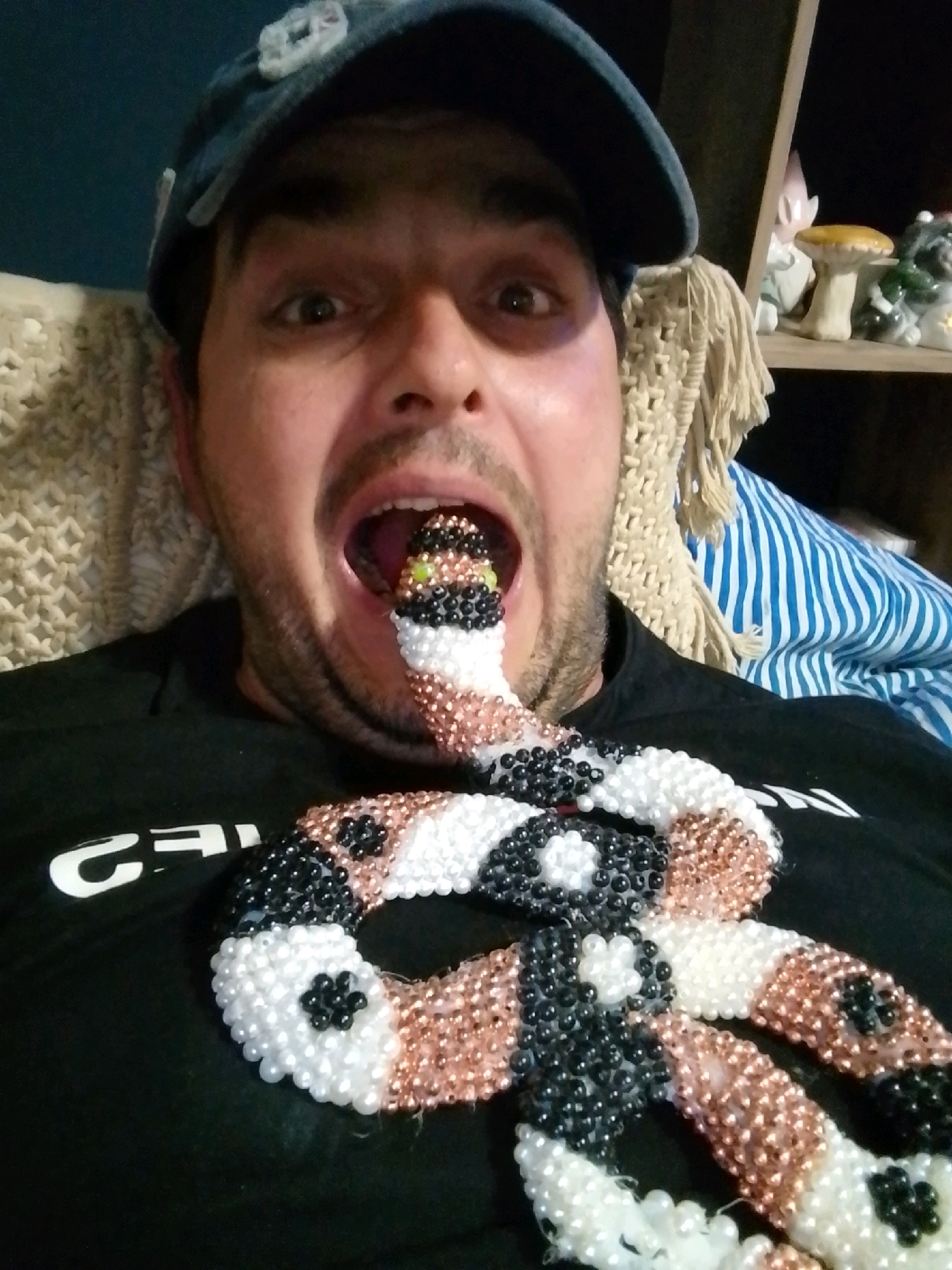 The image shows a man wearing a blue baseball cap and a black t-shirt. He is lying down, with a beaded snake-like object in his mouth. The snake is white, black, and copper-colored, with a patterned design. In the background, there is a beige knitted blanket, a blue and white striped fabric, and a wooden shelf with decorative items, including figurines of gnomes and mushrooms. The man's eyes are wide open, and his mouth is open around the snake. The lighting suggests an indoor environment. The image does not provide any information about the city of Wedel, Germany.