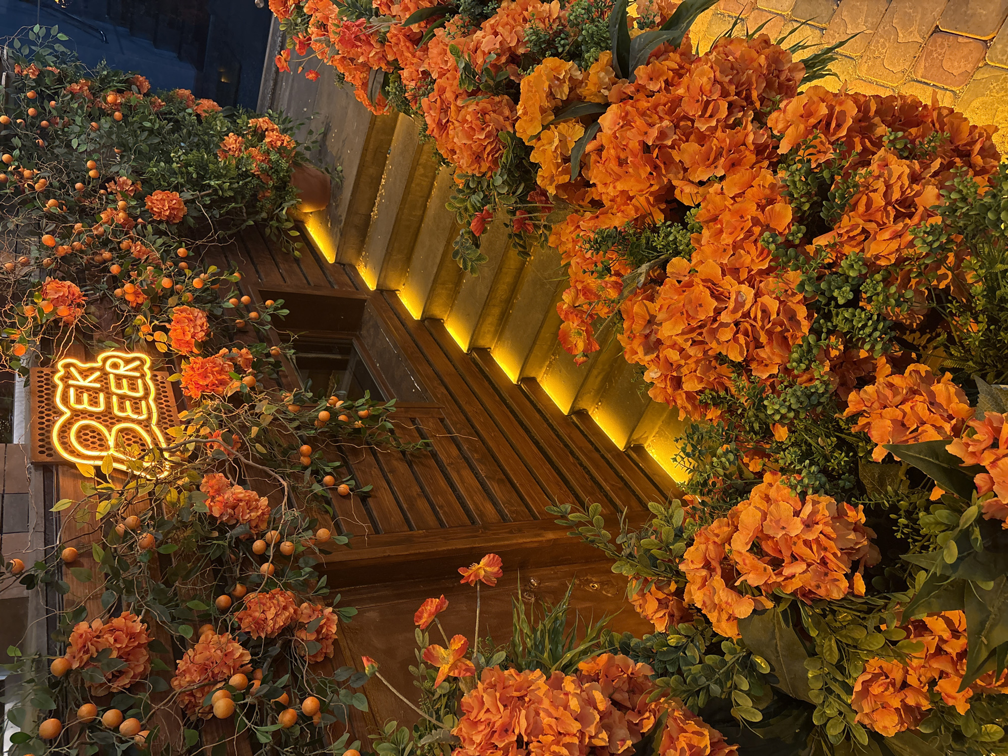 A street-level, close-up, color photograph depicts a building facade adorned with vibrant orange floral decorations and greenery, possibly in Yerevan, Armenia. The facade is primarily constructed of wood and features a distinctive geometric design with angled panels, some illuminated by a strip of warm yellow lighting along their edges. A neon sign with indecipherable writing is mounted on the wooden facade to the left. The floral decorations consist of dense clusters of orange blossoms, resembling hydrangeas, and trailing vines bearing small, round orange fruits, intermixed with green foliage. The composition is arranged to emphasize the abundance and vibrancy of the floral decorations and the distinctive architecture.