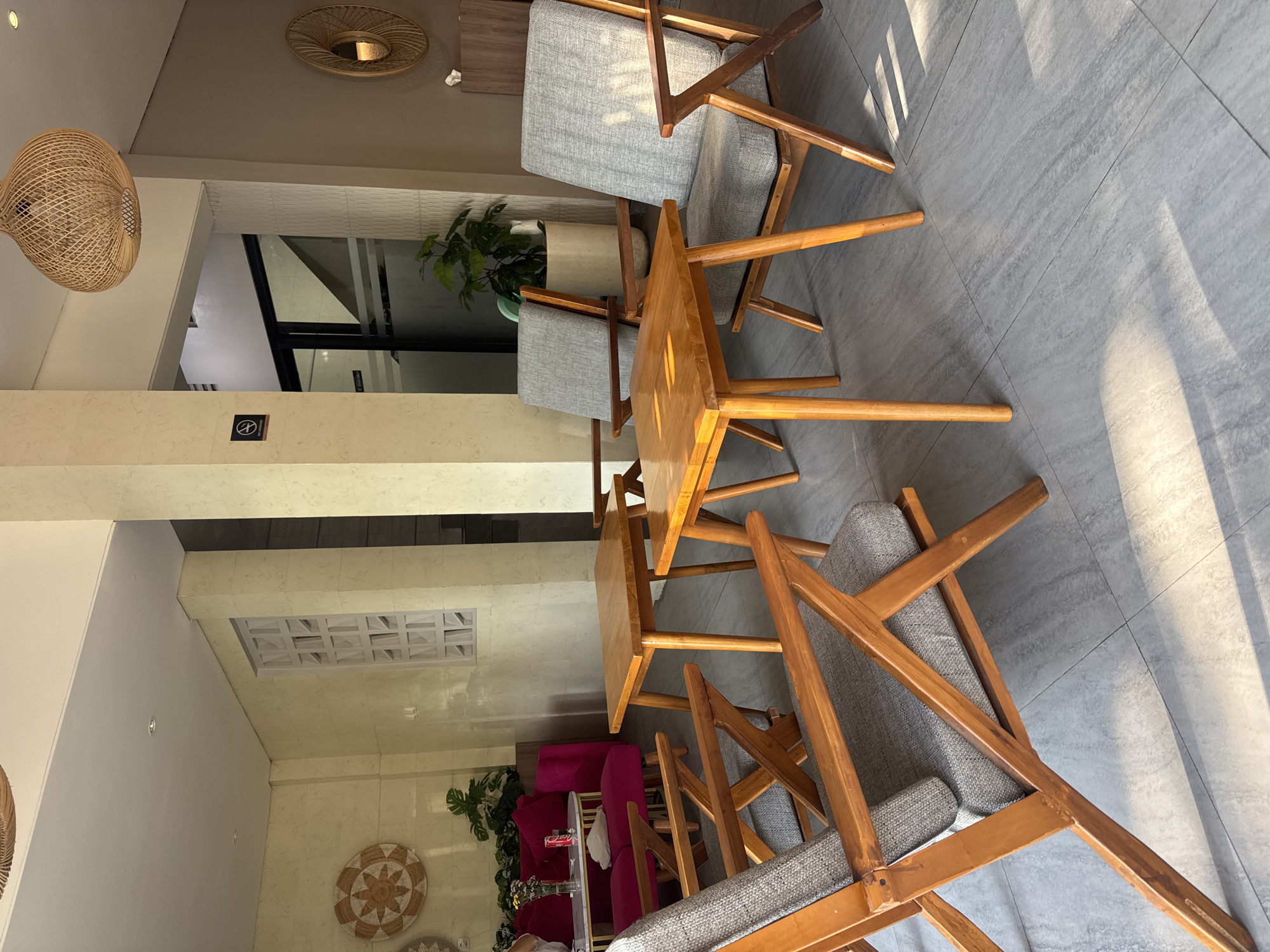 The scene is an interior space in Kuta, Indonesia, featuring wooden chairs with gray cushions arranged around small, square wooden tables. The chairs and tables are positioned on a gray-tiled floor, and the lighting suggests daylight entering from the right side of the frame. In the background, there is a wall decorated with woven discs, along with a magenta sofa and plants. Above, there are rattan-style light fixtures and recessed ceiling details.