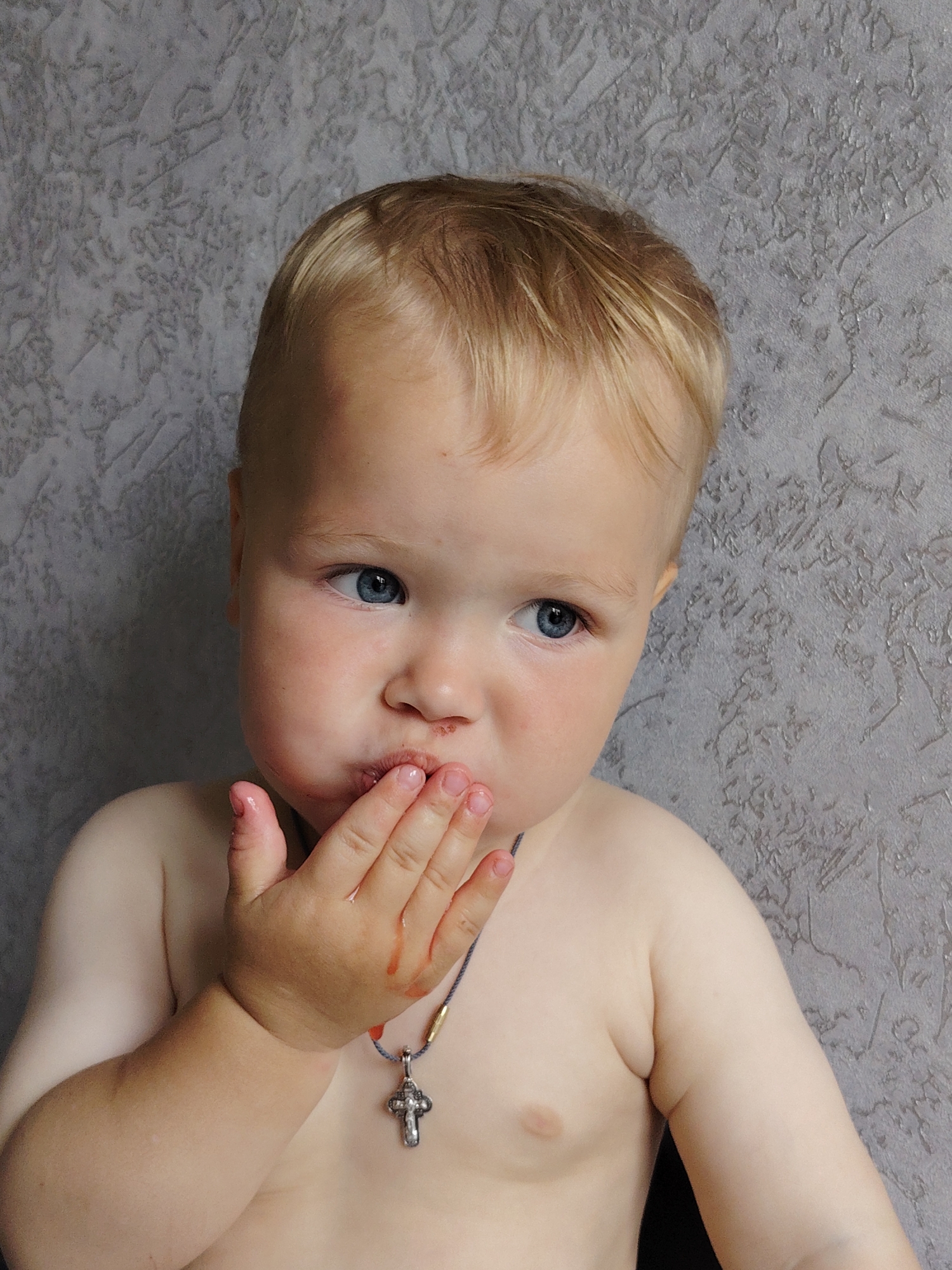 A close-up, chest-up, medium shot captures a fair-skinned toddler with blond hair and blue eyes, posed against a textured gray wall. The child's right hand is placed over their mouth, with their fingers smudged with red, likely from food. A delicate silver cross pendant on a cord hangs around their neck. The toddler's gaze is directed off-camera to the right, with a slightly pinkish hue to their cheeks. The background is a nondescript gray textured wall, indicating an indoor setting. There is no discernible location in the city of Chaikovsky, Russia, depicted in the image.