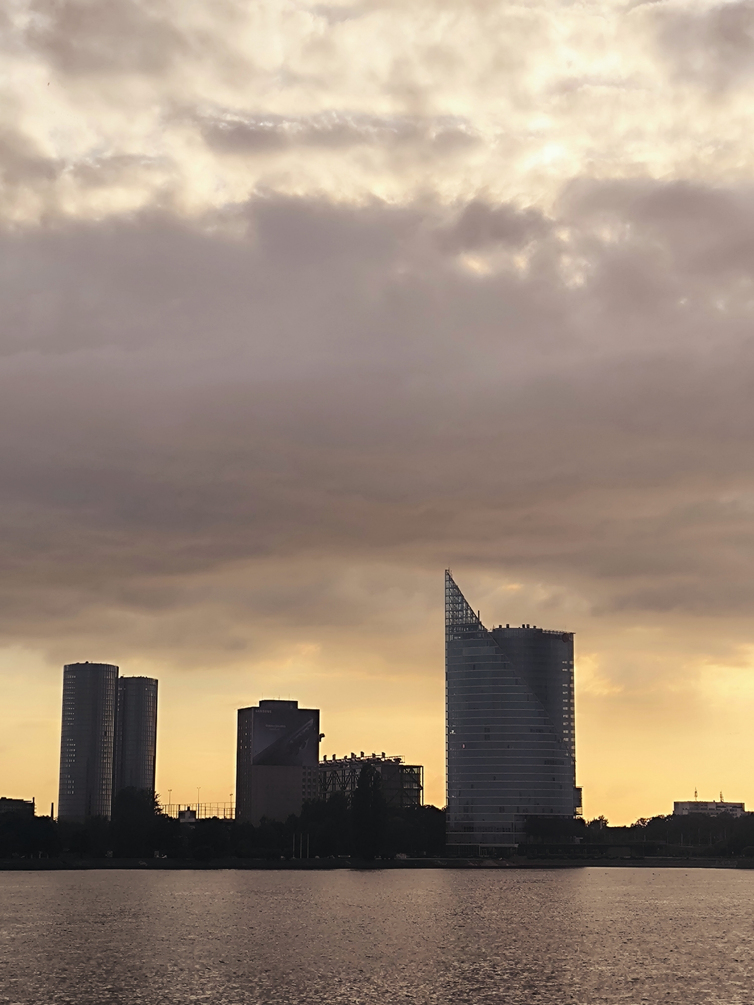The image depicts a view of the Riga skyline from across the Daugava River at sunset. The sky is filled with dramatic clouds, ranging in color from soft yellow and orange near the horizon to deeper purple and gray at the top. Several modern skyscrapers are visible, silhouetted against the colorful sky.
On the left, two tall, slender buildings with many windows stand close together. To their right is a boxier building with a large advertisement displayed on its side. Further to the right, a distinctive, curved skyscraper with a pointed top dominates the scene. This building appears to be under construction or renovation, with scaffolding visible along its upper edge. The base of the buildings is lined with dark trees and other structures, forming a dense urban landscape.
The Daugava River occupies the foreground, its surface rippling and reflecting the subdued light of the sunset, creating a textured, brownish-gray expanse. A few distant, small birds are visible in the sky. The overall impression is of an urban waterfront at dusk, with the city's architecture set against a vibrant, cloudy sky.
