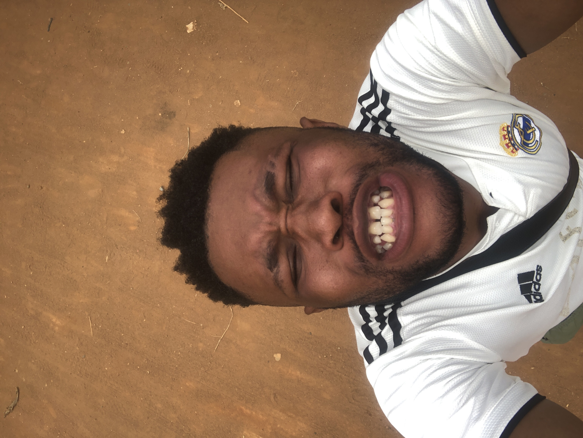 A man with brown skin, dark hair, and a beard is seen wearing a white t-shirt with black stripes and a Real Madrid logo on the chest. He is grimacing with his eyes closed and teeth bared. He is lying on the ground, which is brown and possibly dirt or sand. The location is unknown.