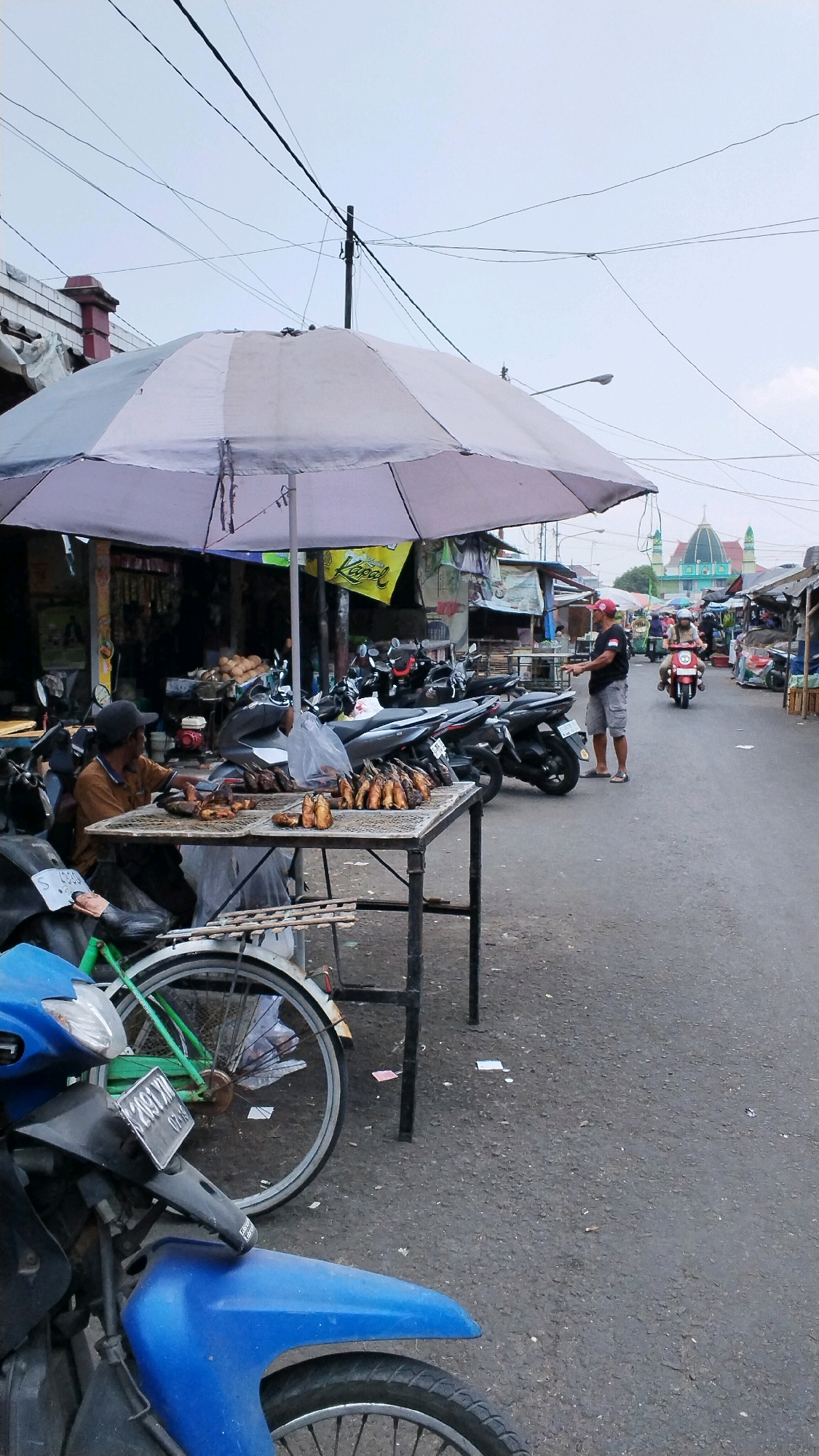 This is a medium shot, eye-level, outdoor, full-frame photograph taken at the Jagalan market in Indonesia. The scene depicts a street market setting, likely in the morning or late afternoon, judging by the overcast sky and diffuse lighting. In the foreground, a blue and black motorcycle is partially visible, parked on the right side of the frame. To the left and slightly behind it, the rear wheel of a green bicycle with a black tire is visible.
Dominating the center of the frame is a makeshift market stall covered by a large, light-colored umbrella. The stall is set up on a table made of metal tubing and wood, displaying a variety of cooked fish. A man wearing a brown shirt and a black cap sits behind the table, appearing to be tending to the stall. He is facing the viewer's left.
Behind the stall, several motorcycles are parked in a row, indicating a common mode of transportation for vendors and customers. The market extends further down the street, with various stalls and buildings lining both sides. In the background, a distinctive building with a green dome and two minarets, likely a mosque, is visible, providing a geographical marker. Another man wearing a red cap and grey shorts stands near the motorcycles, looking towards the market. The street is paved with asphalt and appears to be a busy thoroughfare for market activity. The overall impression is one of a lively, local marketplace.