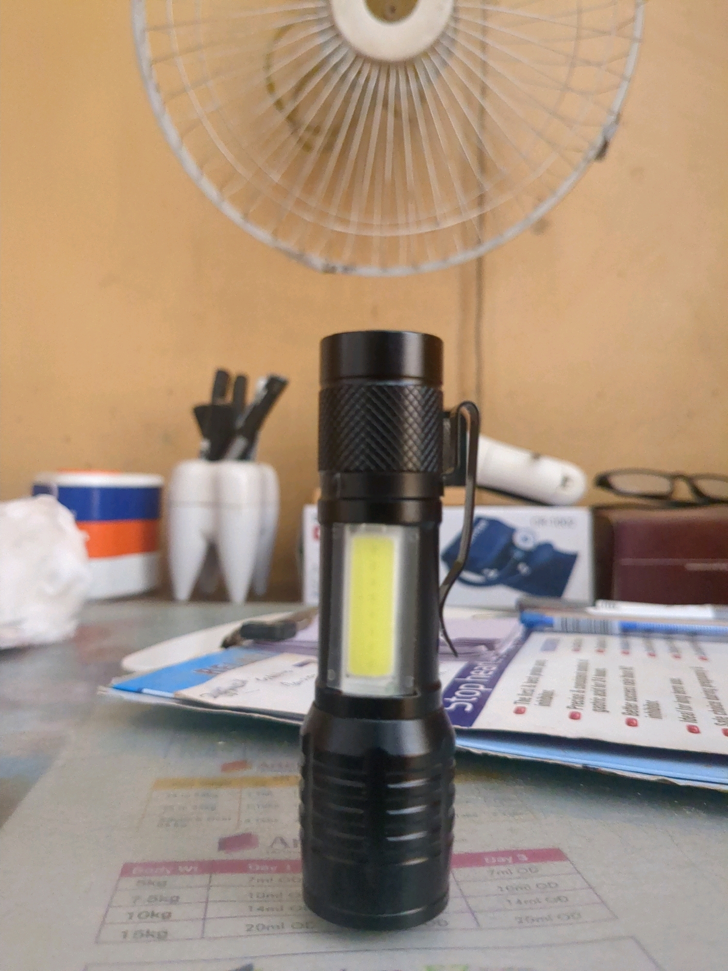 The image shows a close-up, eye-level shot of a black LED flashlight with a white LED light strip positioned vertically. The flashlight is on a table, with a blurred background. To the left of the flashlight, there is a white container shaped like a tooth, holding several black pens or markers. Further left, there is a cylindrical object with blue and orange bands, possibly a container. In the background, a white oscillating fan with visible blades is mounted above, and a wall with a textured, light brown surface is visible. The table surface also displays some papers with text and what appears to be a medical chart with measurements. A pair of glasses and a dark brown rectangular object are also visible in the background, to the right of the flashlight. The overall scene suggests an indoor setting, possibly an office or study. There are no people visible in the image. The image does not provide any specific geographical or locational information that would place it within Karachi Division, Pakistan.