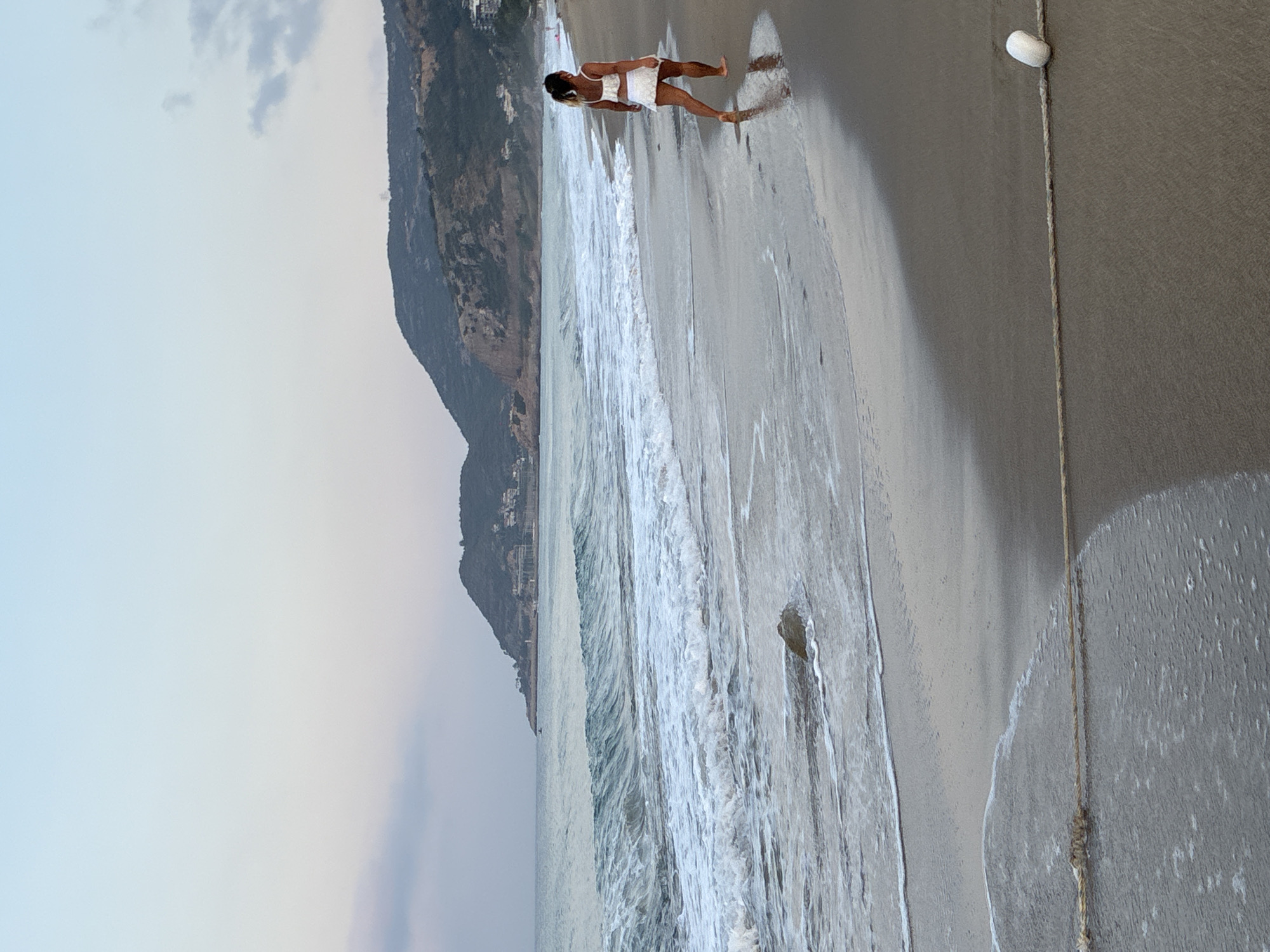 A lone woman in a white top and skirt walks along a wet sandy beach towards the right of the frame. The woman is facing away from the camera and appears to be walking along the shoreline. The sea, with moderate waves, is to her left. In the background, a hilly coastline with buildings and trees is visible. The sky is a soft gradient of pale blue and pink, suggesting either sunrise or sunset. The scene is set in Alanya, Turkey, likely on one of its popular beaches. A rope with a white float is partially visible in the foreground, running horizontally across the sand.