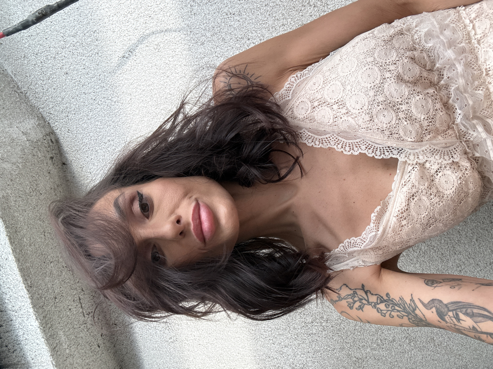 A woman lies on a light grey, textured surface. She wears a light-colored, lace-trimmed top. She has dark hair, brown eyes, and pink lips. Tattoos are visible on her left arm and shoulder. The image does not contain sufficient information to determine a location in Pleven, Bulgaria.