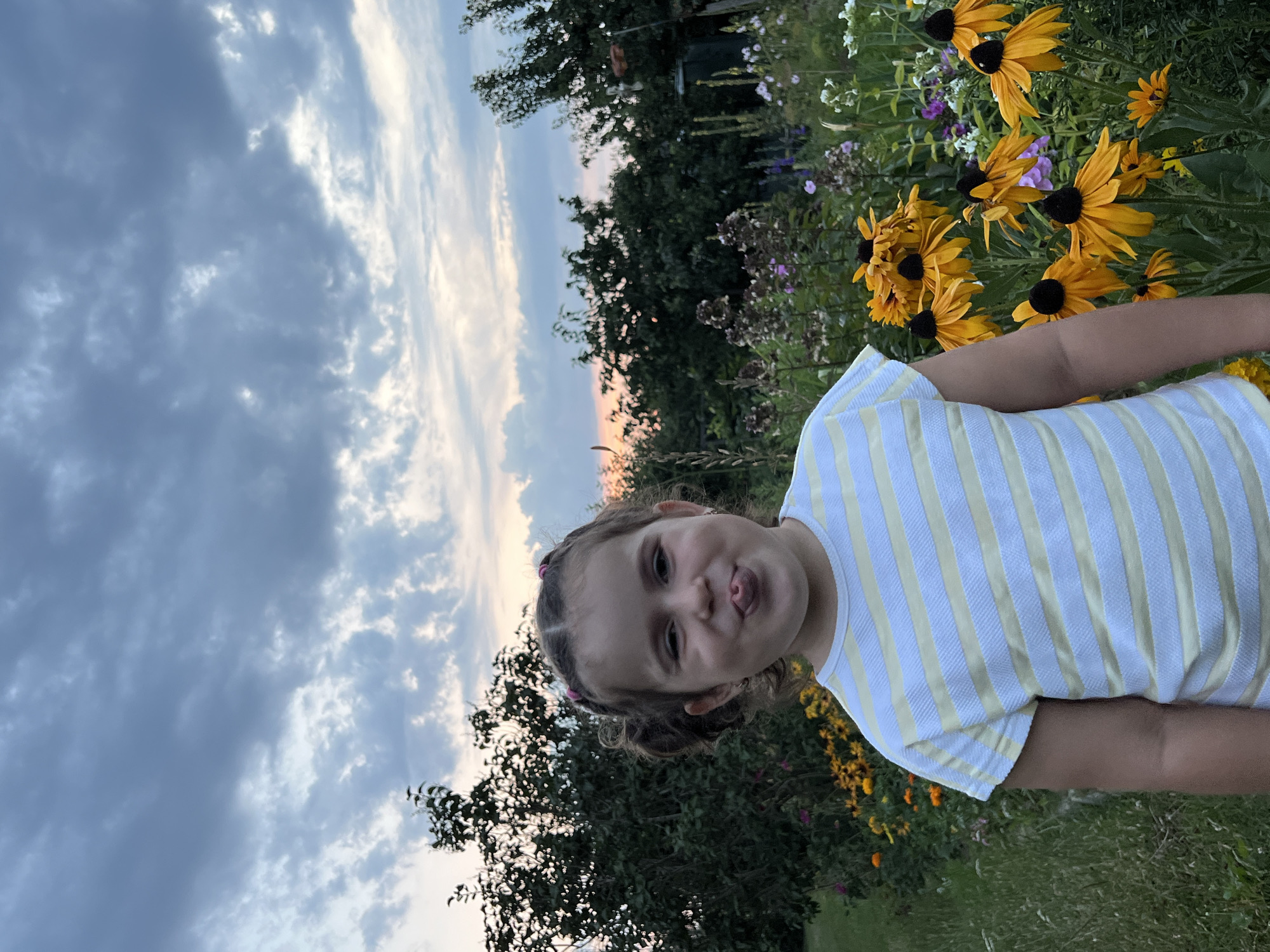 A young girl with her tongue sticking out stands in front of a garden. She has brown hair pulled into two pigtails. She is wearing a yellow and white striped t-shirt. The garden is filled with yellow and purple flowers and green foliage. In the background, there are trees and a cloudy sky with hints of sunset colors. The location is Elizówka, Poland.