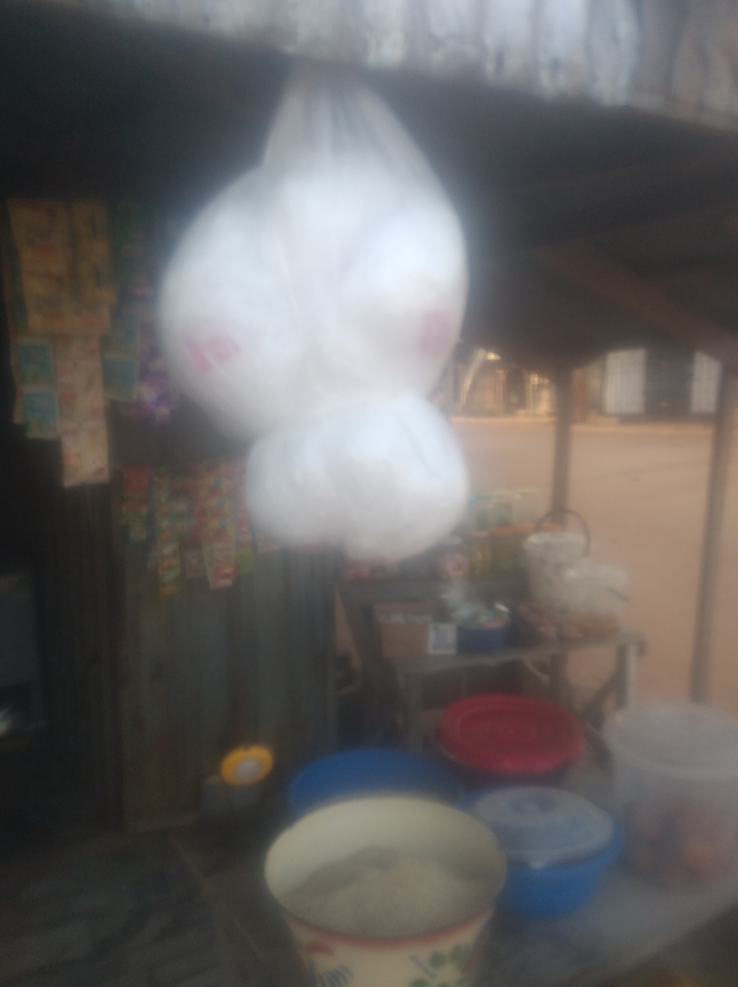 The image depicts a roadside stall in Jalingo, Nigeria. Two large, translucent white plastic bags, likely containing merchandise, are hanging from an overhead structure. Below them, a shelf displays various items including what appears to be packaged goods and containers. On the ground level, several plastic containers of different colors are visible, some with lids. The stall is set up against a backdrop that suggests an outdoor urban environment, possibly a street market. The overall impression is one of a typical informal commercial setup in Nigeria.