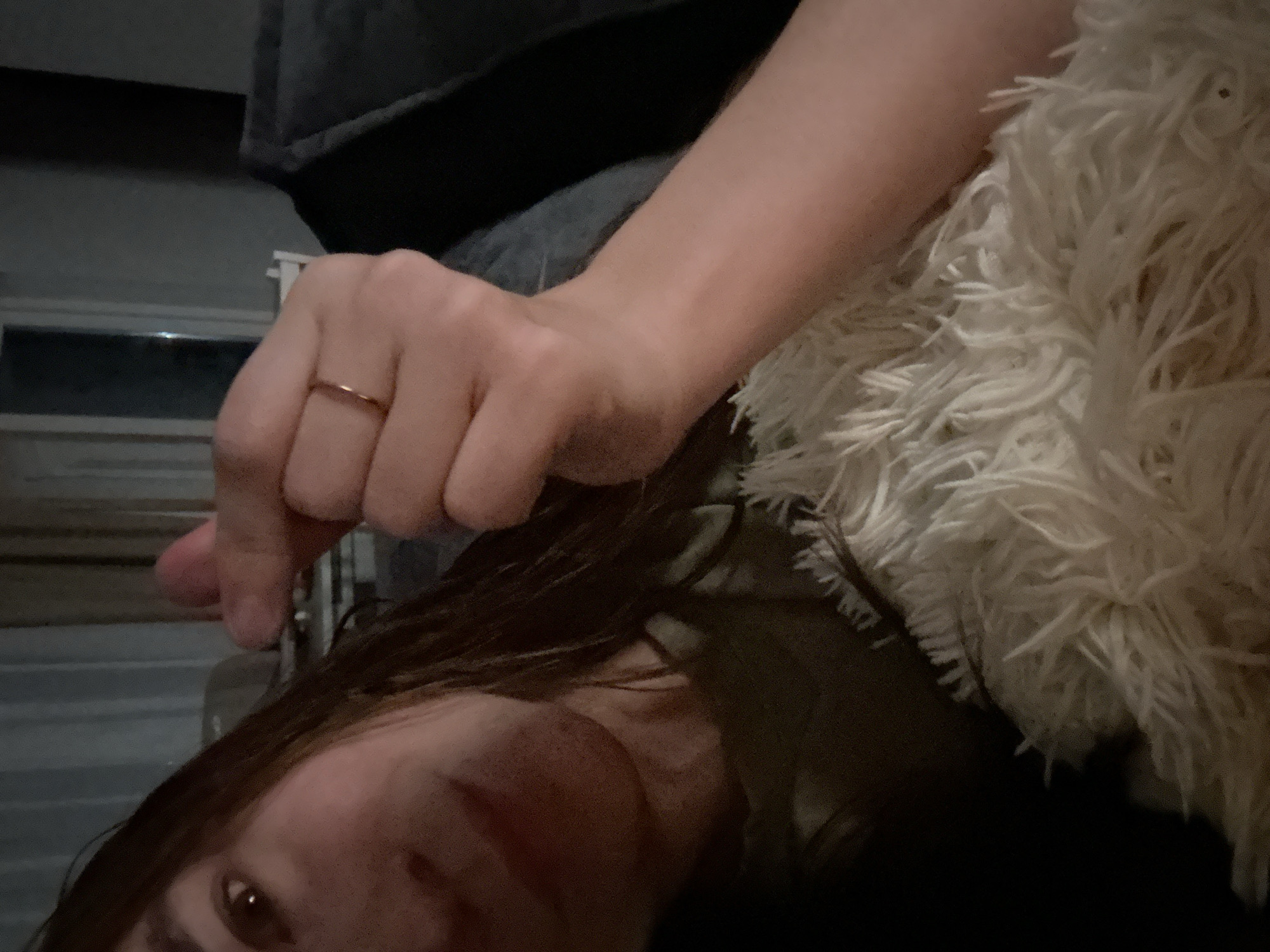 The image captures a close-up, angled view of a person lying down indoors in Kharnavy, Russia, under dim lighting, suggesting evening or nighttime. The lower left features a partial view of a face with brown hair, including an eye, nose, and mouth. An arm extends into the upper left, with the hand forming a "finger heart" gesture, indicating affection. A simple, thin gold-colored ring is visible on the person's ring finger. A fluffy, cream-colored blanket or pillow occupies the right side of the frame, partially covering the subject. Dark furniture or bedding is in the background on the upper left, with a faintly visible window or reflective surface showing horizontal lines. No text is visible.