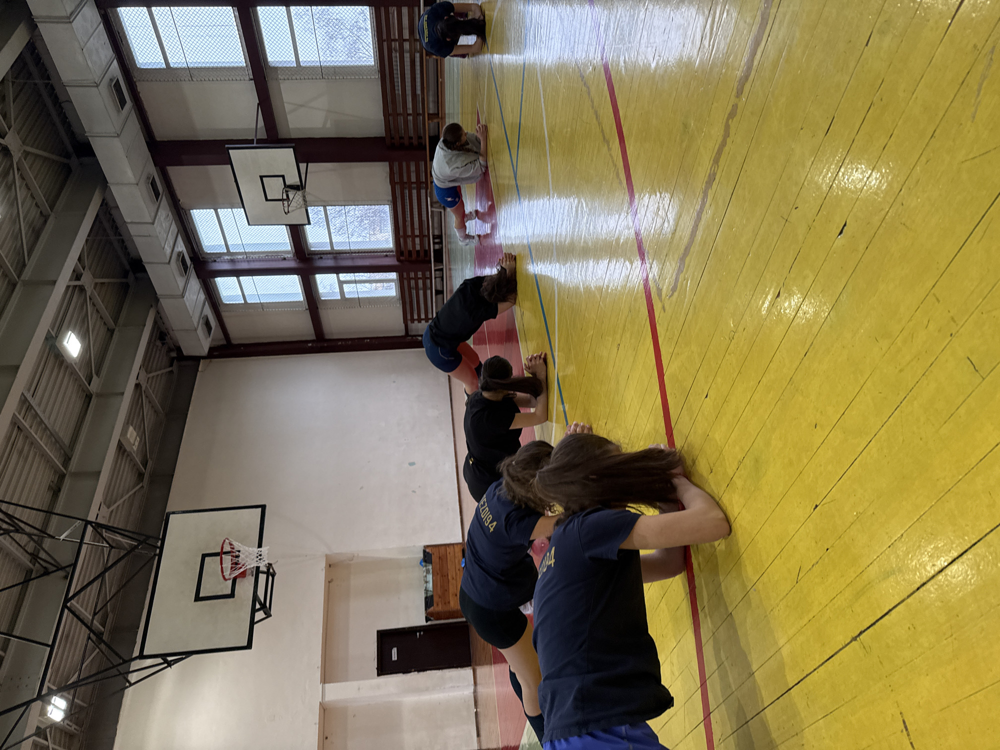 The image displays the interior of a sports hall located in Sofia, Bulgaria. The specific name or address of the facility cannot be ascertained from the visual evidence. Seven individuals, identified as female, are performing a synchronized forearm plank exercise on a glossy, yellow wooden floor marked with red and blue court lines. The majority of participants wear dark blue or black athletic t-shirts and shorts; one individual in the mid-ground wears a light gray long-sleeved top and blue shorts. The floor exhibits signs of wear and scuff marks.
The environment includes light-colored walls. On the left, a dark brown door is visible above a wooden crate-like object. Along the right wall, multiple tall, rectangular windows with internal wire mesh or grates allow daylight to enter, and bare tree branches are discernible outside. Horizontal wooden benches or bleachers are mounted on the wall beneath these windows. The high ceiling features exposed metal framework, light gray rectangular ventilation ducts, and rectangular fluorescent light fixtures. Two basketball hoops, each with a white backboard and a red-and-white net, are mounted on the walls; one is prominent on the left and another is partially visible on the right.