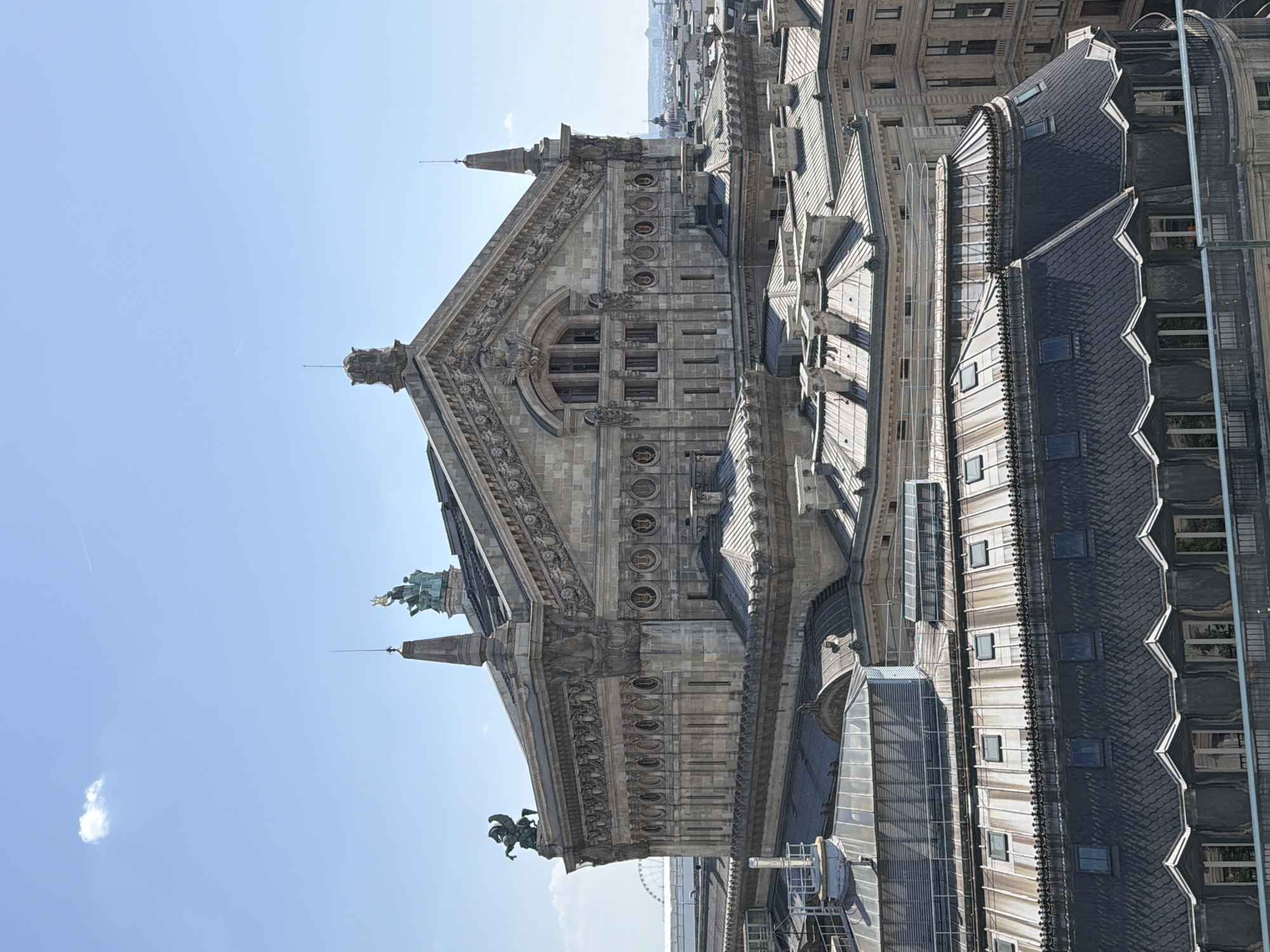 The image, taken from a high vantage point in Paris, France, captures a detailed view of an ornate, stone building and its surrounding rooftops under a clear blue sky. The building's architecture is characterized by intricate carvings, decorative friezes, and multiple levels of windows. A prominent feature is a large, triangular pediment adorned with sculptures and architectural ornamentation. Statues of figures, possibly mythological or historical, are visible on the building's cornices.
The scene is illuminated by bright sunlight, suggesting it's daytime, likely midday given the strong shadows. A faint airplane contrail streaks across the sky, and a small, wispy cloud is visible in the upper left quadrant. Adjacent to the main building, a series of dark, tiled roofs with numerous skylights stretch into the distance, showcasing a dense urban landscape typical of Paris. The perspective offers a sense of scale, emphasizing the grandeur of the architecture against the expansive sky. No specific activity or event is depicted; the focus is on the architectural details and the urban setting. There is no visible text in the image.