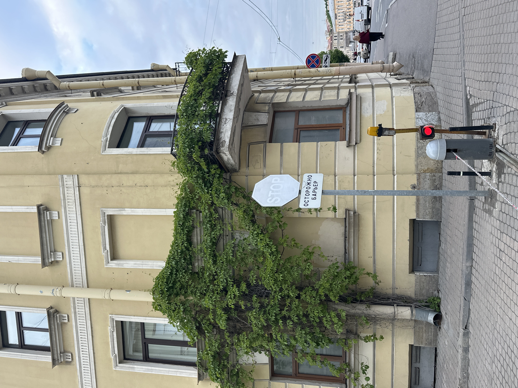 The image shows a street scene in Saint Petersburg, Russia. A yellow building with decorative window frames and a wrought-iron balcony is partially covered in green ivy. A white, octagonal "STOP" sign and a rectangular sign with Cyrillic text ("ОСТОРОЖНО БАРЬЕР," meaning "Caution Barrier") are mounted on a pole attached to the building. To the right, a traffic light shows a red signal. A grey mechanical barrier device with a white dome and attached chain sits on the sidewalk. In the background, a street with parked cars and a few pedestrians is visible. The sky is overcast. The image is rotated, so the scene appears sideways.