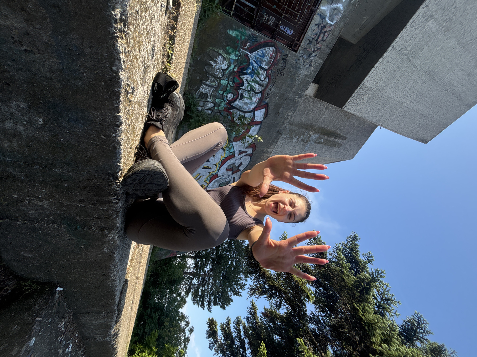 This dynamic image captures a young woman in an unusual, rotated perspective, striking a playful and energetic pose outdoors. The photograph is oriented 90 degrees clockwise, making the woman appear vertical along the left edge of the frame, as if she is leaning against a wall or suspended. She wears a dark grey/brown ribbed tank top, matching leggings, and black athletic shoes. Her hands are extended towards the camera with fingers spread wide, and her mouth is open in a wide, joyful, or excited expression, suggesting a fun interaction or a dramatic pose.
The setting appears to be an urban or semi-urban environment in Varna, Bulgaria. She is positioned against a rugged concrete surface that forms a substantial ledge or wall. Behind her, a large concrete structure is extensively covered in colorful graffiti, adding a vibrant, artistic, and somewhat gritty backdrop. Visible text within the graffiti includes "ODOD" and possibly "DARNA TROP". Below the main concrete structure, dense green coniferous trees provide a natural element. The upper right portion of the image features a clear, bright blue sky, indicating a sunny day. The low camera angle enhances the sense of scale and drama, creating a unique visual experience. The lighting suggests it is daytime, with clear weather.