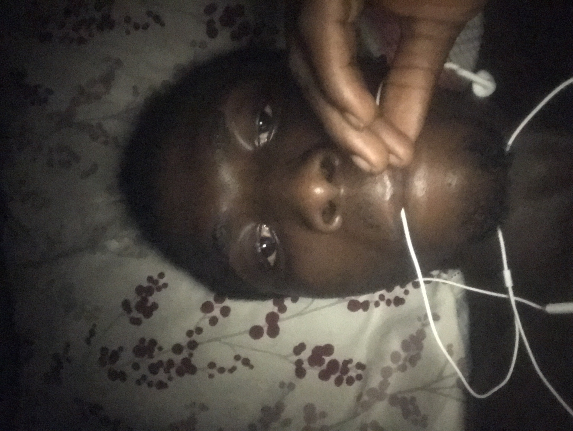 A dark-skinned person is lying down on a patterned sheet, with their head and upper body filling the frame. The pattern on the sheet appears to be small, dark red circles scattered on a light background, resembling a floral or berry design. The person is looking directly at the camera, with their right hand near their mouth, holding something white that appears to be a part of earbuds. White earbuds and wires are visible around their neck and on the sheet. The lighting is dim, creating shadows and highlighting certain features of the face. The image has a grainy quality.