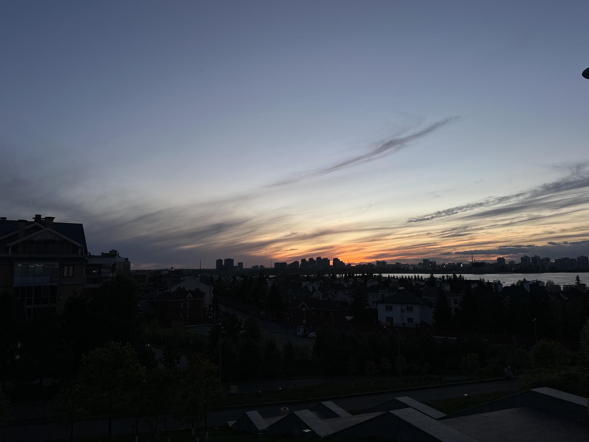 The image depicts a sunset view of Kazan, Russia, with a focus on the cityscape and its reflection on a body of water, likely the Kazanka River or Lake Kaban. The foreground features residential buildings and trees silhouetted against the fading light. In the distance, the Kazan skyline is visible, marked by high-rise buildings. The sky displays a gradient of colors, transitioning from dark blues and purples at the top to orange and yellow hues near the horizon. Clouds are present, adding texture to the sky. The image appears to be taken from an elevated position.