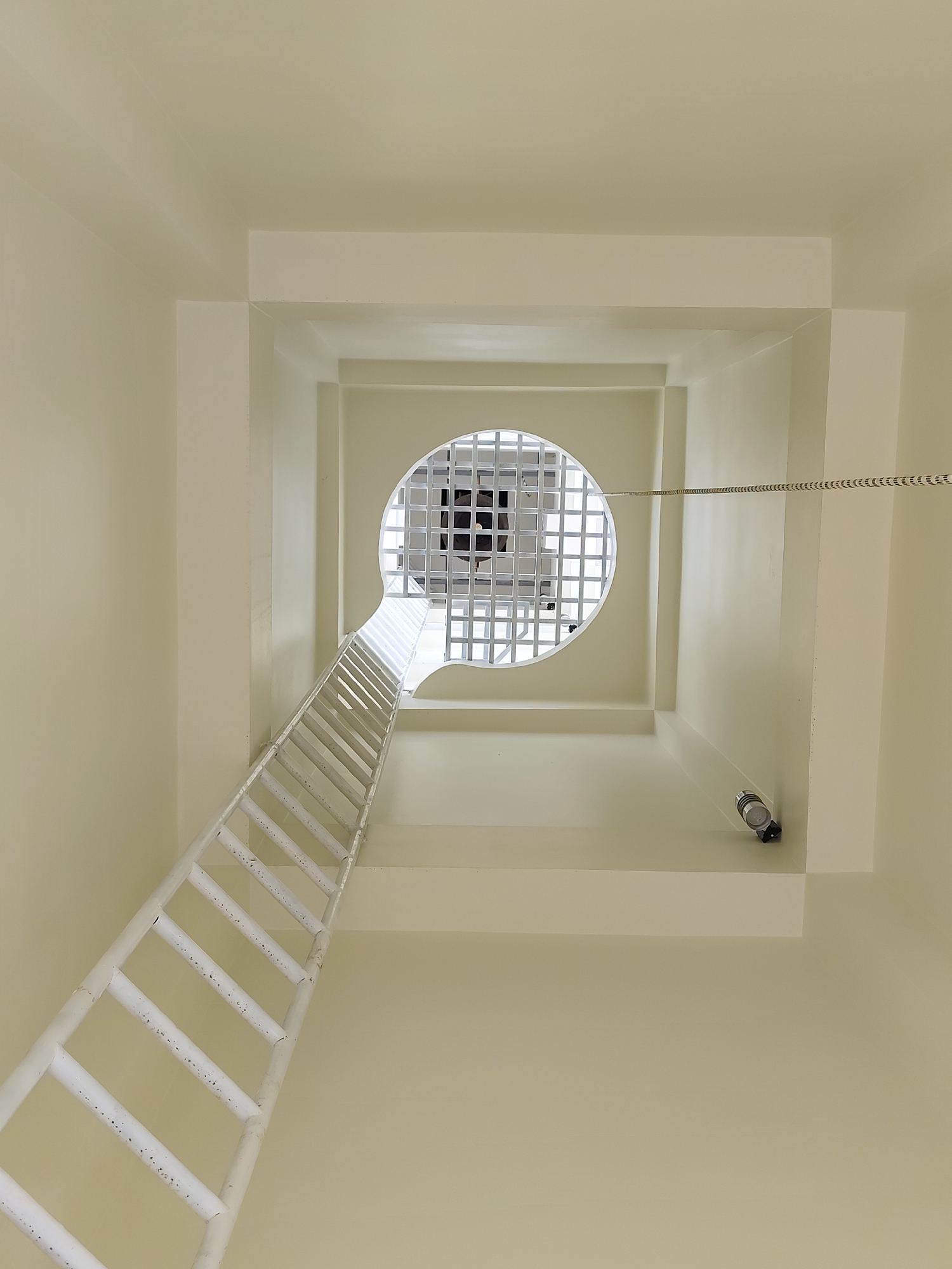 Looking upwards, the image shows a tall, narrow vertical shaft painted in a pale cream color. A white metal ladder extends from the bottom of the frame, appearing to ascend towards a circular opening at the top. Within the circular opening, a grid-like metal structure is visible against a bright, possibly outdoor, backdrop. A cable or chain stretches horizontally across the shaft, parallel to one of the upper sections. A small cylindrical object, possibly a light fixture, is mounted on the wall near the upper right side of the frame. The location is specified as Đa Thọ, Vietnam.