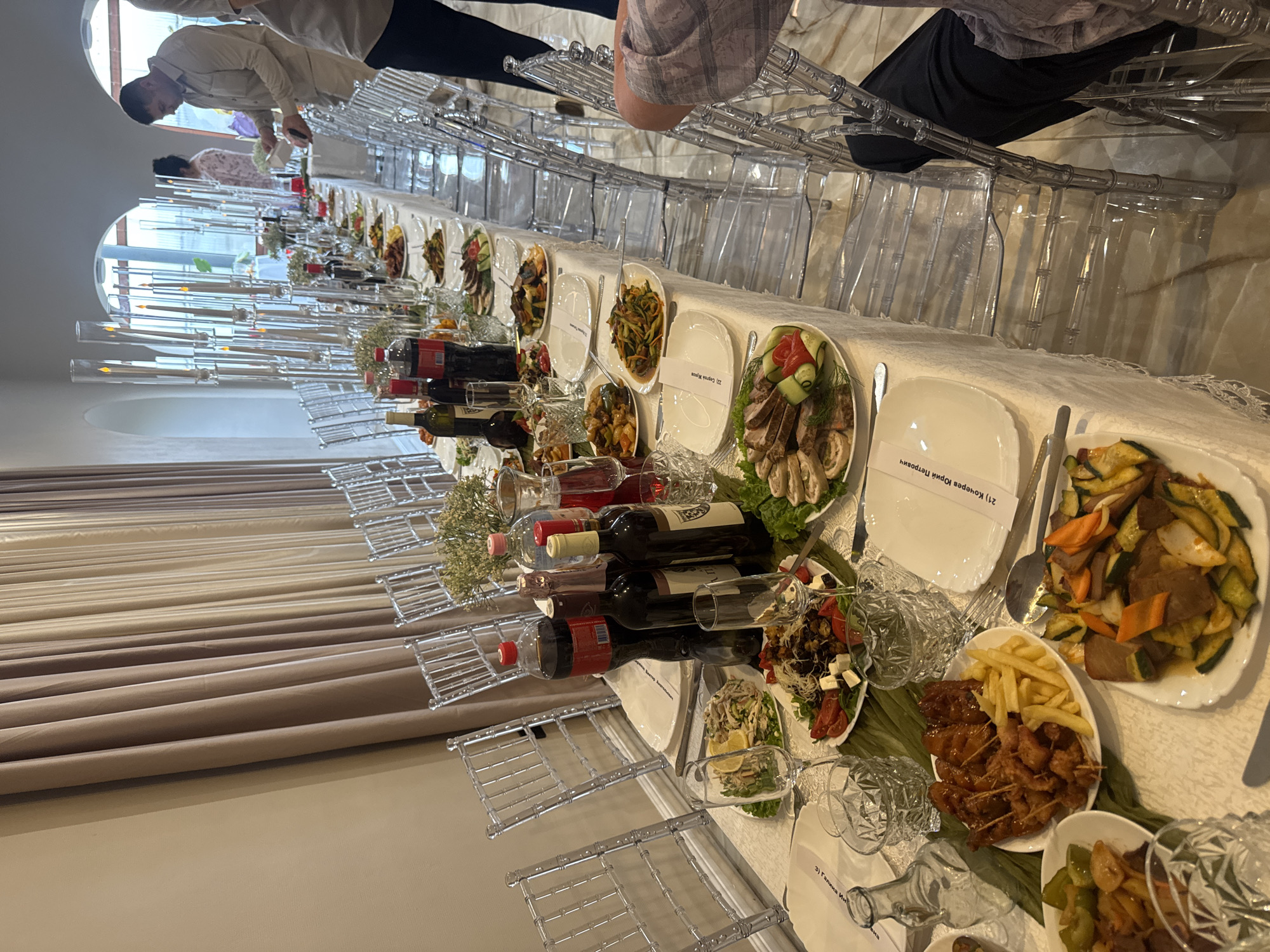The image depicts a banquet table set for a formal event, likely a wedding reception or significant celebration, in Komsomolsk-on-Amur, Russia. The table is adorned with white linen, multiple plates of assorted appetizers and main courses, bottles of wine and soda, and decorative glassware. Transparent chairs are arranged along the sides of the table, with some occupied by guests. In the background, a large window or glass-paned wall reveals what appears to be an outdoor area. The lighting is dim, suggesting an evening event. The overall scene conveys an atmosphere of festivity and abundance.
