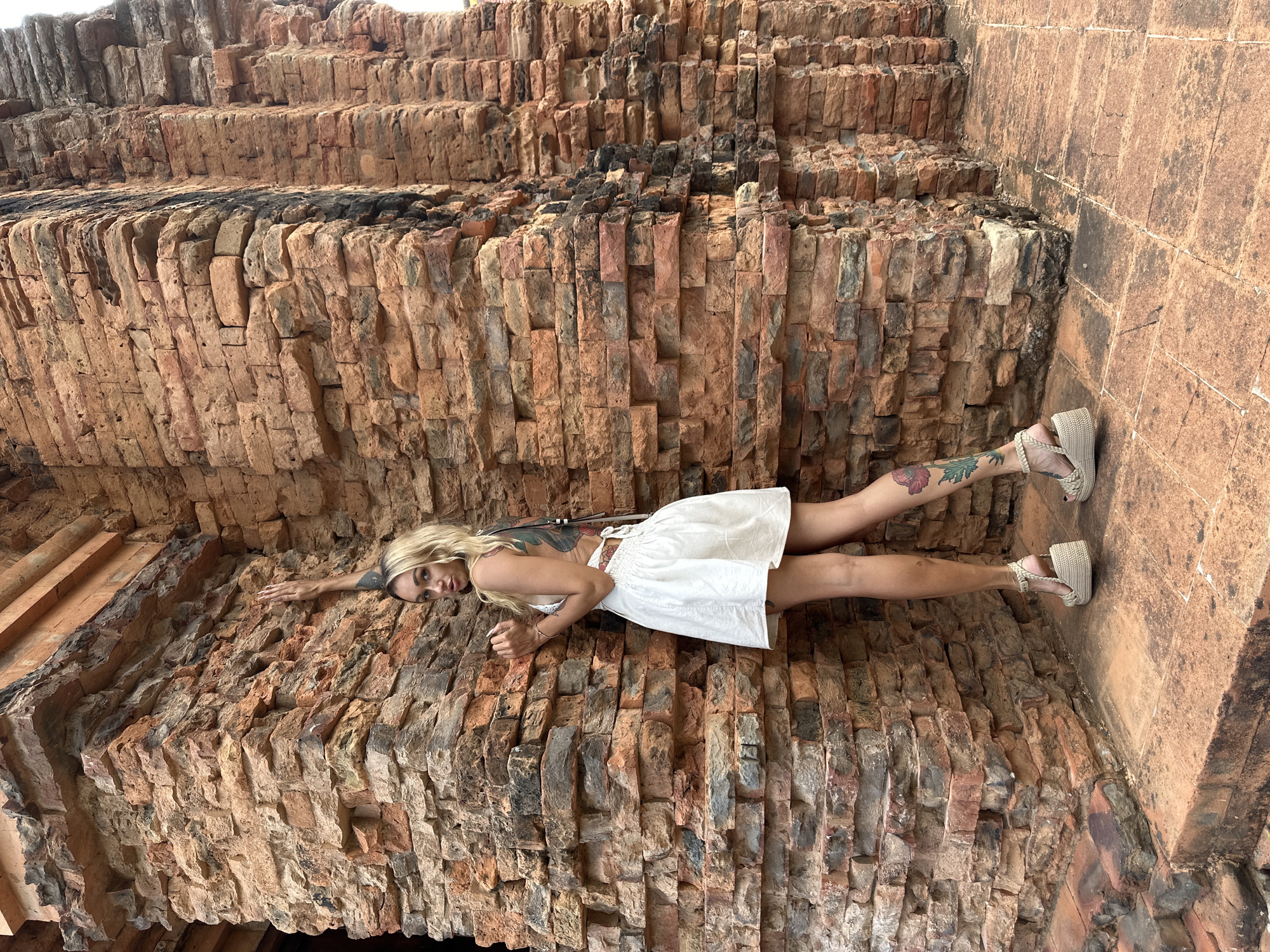 A woman lies on a brick structure in Phu Thuy Ward, Vietnam. The woman, fair-skinned and blonde, wears a short white dress and braided sandals. She has tattoos on her right leg and back. Her head is near the bottom left of the frame, with her body extending diagonally across the image to the top right. The backdrop consists of aged, reddish-brown bricks arranged in layers.