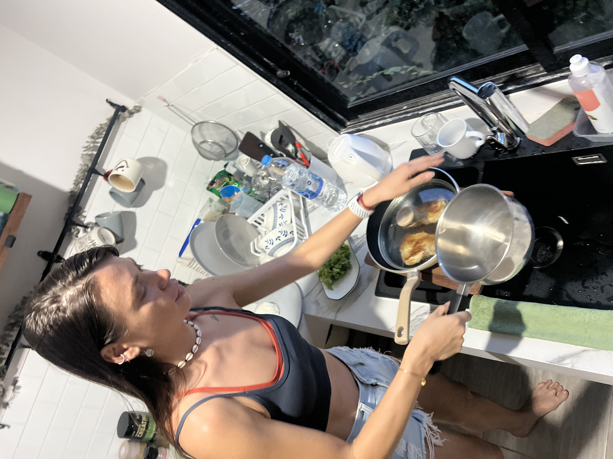A woman is in a kitchen in บ้านบางรักษ์, Thailand. She is barefoot, wearing denim shorts and a dark blue top with red trim, and a shell necklace. She is cooking at a stove, pouring liquid from a metal saucepan into a frying pan containing food. A window is above the sink, next to which are various cups, containers and cleaning supplies. Several dishes and kitchen utensils are stacked near a bottle of water.