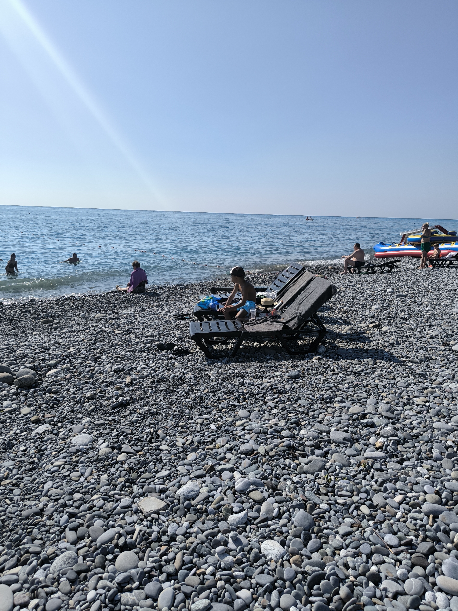 A sunny day at a pebble beach in Sochi, Russia. The clear blue sky is vast above the tranquil sea. People are scattered across the beach and in the water. A young boy in blue shorts sits on a dark-colored lounge chair on the left side of the frame, facing the sea. Next to him, another lounge chair is occupied by a person. Further back, near the waterline, a woman in a purple cover-up sits on the pebbles, facing away from the sea. Several individuals are wading or swimming in the shallow water, marked by buoys. In the background, inflatable watercraft and a few distant boats are visible on the sea. The beach itself is covered in a dense layer of smooth, dark gray pebbles.
