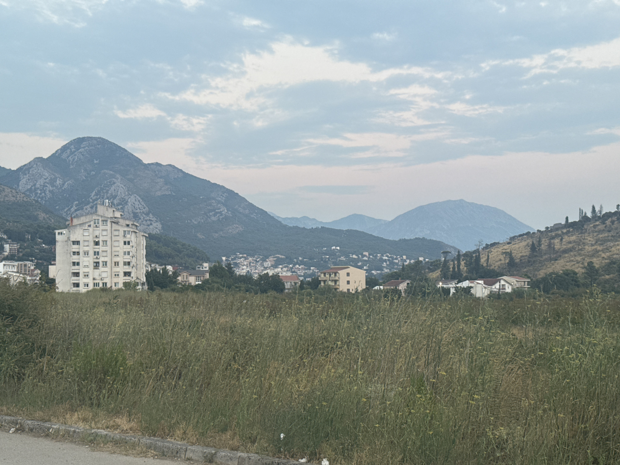 The image captures a view of Sutomore, Montenegro, dominated by a large, multi-story, light-colored apartment building with numerous windows and balconies. In the foreground, a dense field of tall, dry grass stretches across the frame, with a paved edge and a curb visible at the bottom left. Behind the apartment building, a town with red-tiled roofs and smaller houses is nestled into the hillside, partially obscured by trees. Further in the background, several layers of mountains rise, with the closest slopes covered in vegetation and the more distant ones appearing as hazy, blue silhouettes against a cloudy sky. The sky itself is overcast with scattered, light gray clouds. The overall impression is a semi-urban landscape with a strong natural backdrop.