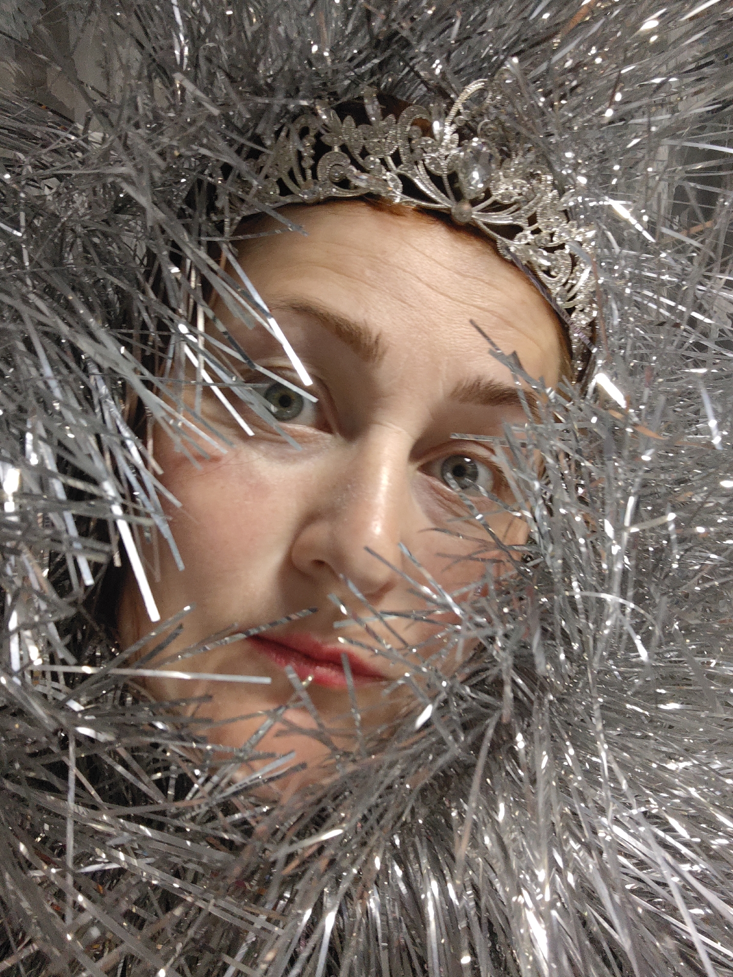 A close-up photograph shows a person with fair skin, light green eyes, and red lipstick, partially obscured by strands of metallic silver tinsel. The person is wearing a silver crown. The location is unknown and there is no evidence to suggest the location is in Марковский, Russia.
