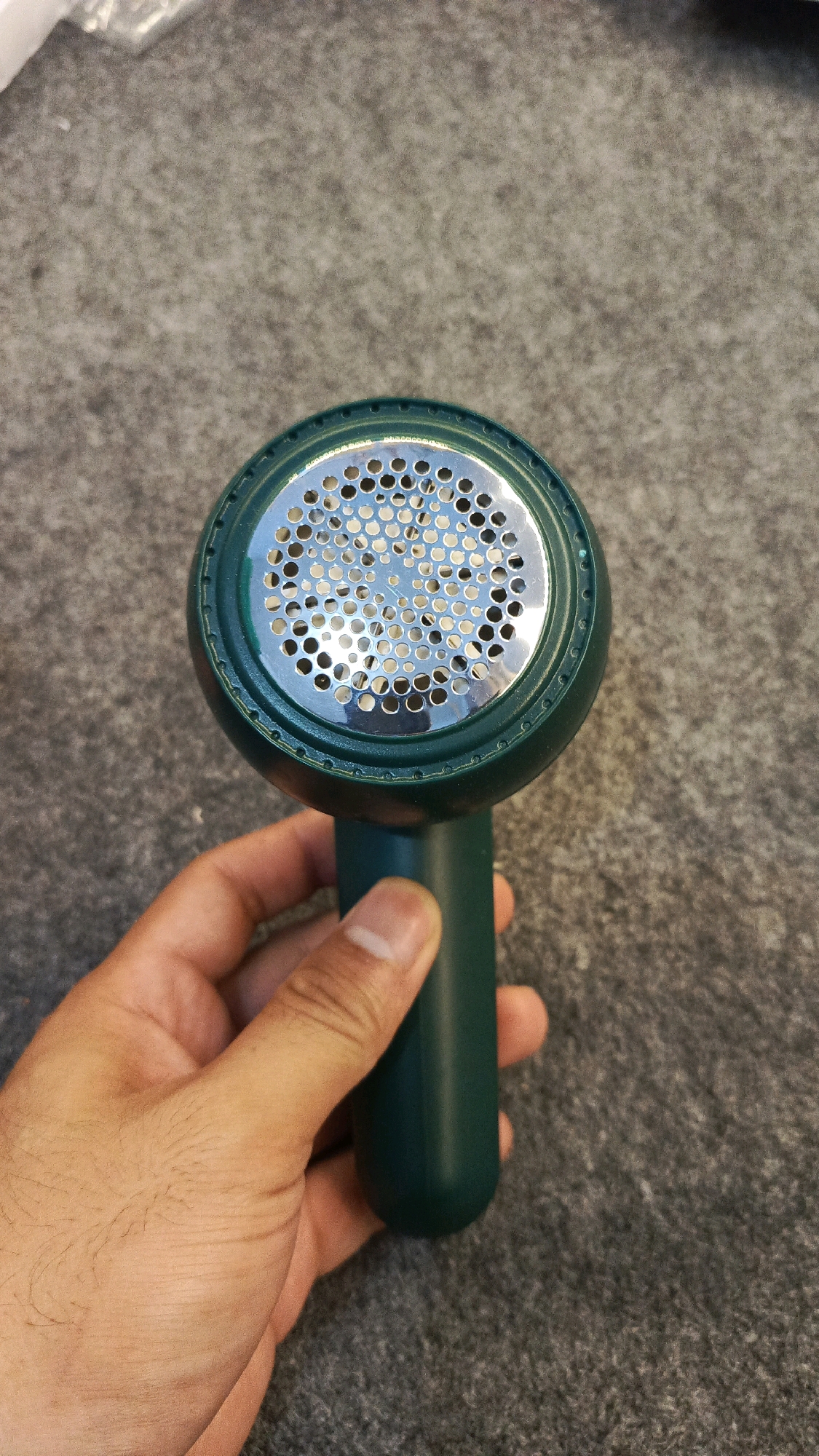 A dark green fabric shaver is held in a person's right hand against a gray, textured surface. The shaver has a circular head with a metallic grille containing many small holes. The handle of the shaver is also dark green and rounded. The person's hand is visible gripping the handle, with fingers and thumb positioned around it. The background is out of focus, showing a consistent gray texture. No elements suggest the location is in Babadan, Indonesia.