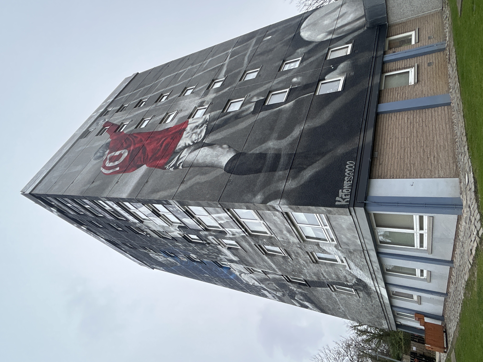 The image shows a tall building in Aberdeen City, United Kingdom, viewed from a low angle. The building's facade features a large-scale mural depicting a person wearing a red jersey with a prominent number 10, set against a grayscale background that includes what appears to be a depiction of the moon. Numerous windows are integrated into the mural design. The building adjoins another structure with a brick-like exterior. Patches of green grass are visible at the base of the building. Graffiti or a signature "KeJones2020" is visible on the painted section of the building. The sky overhead is overcast.